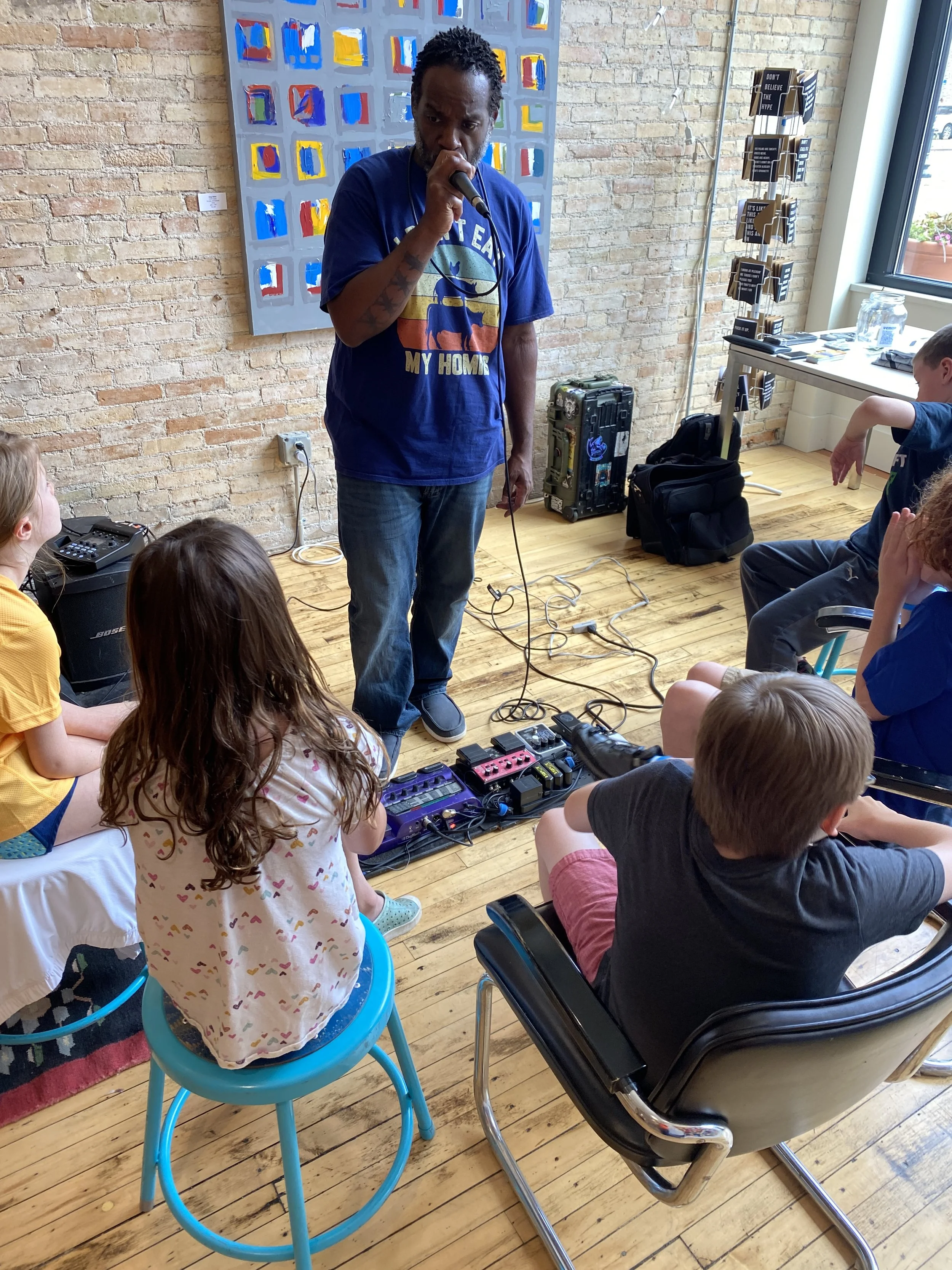 A man with black curly hair and a beard is standing in front of a group of children, holding a microphone and speaking. The man is wearing a blue T-shirt with a graphic and text, and there is a colorful abstract painting on the brick wall behind him.