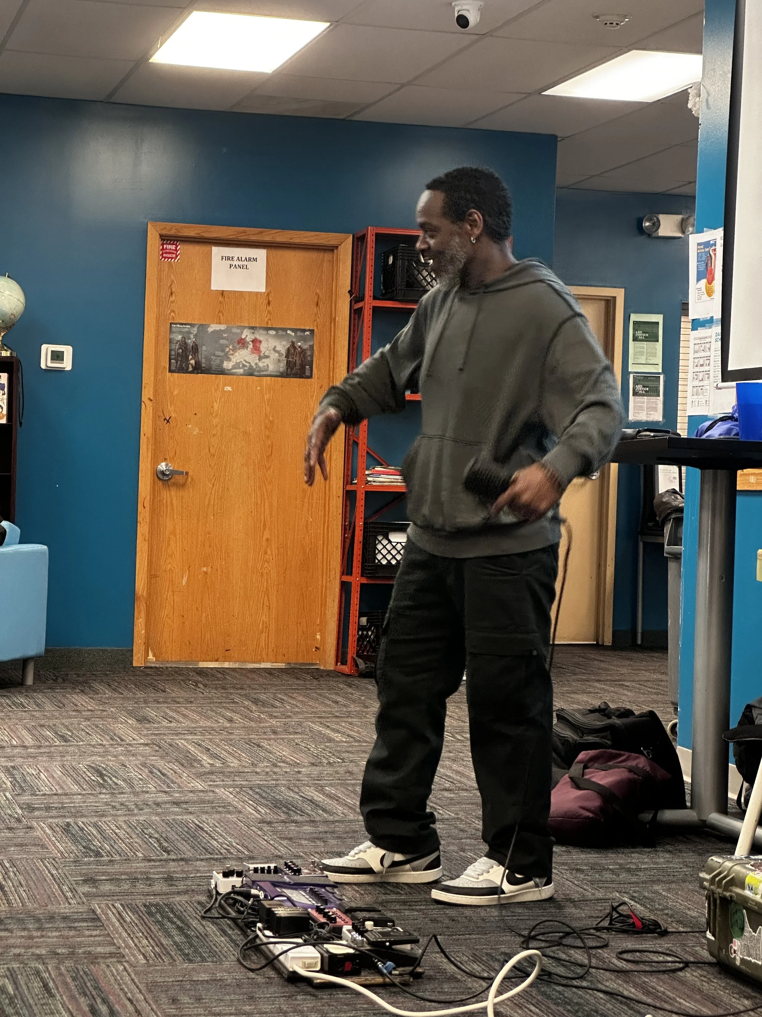 A man with dark hair and a beard, wearing a gray hoodie and black pants, stands on a patterned carpeted floor in an indoor setting He is in front of a pedal board with various effects pedals connected by cables. There is a maroon backpack on the grou