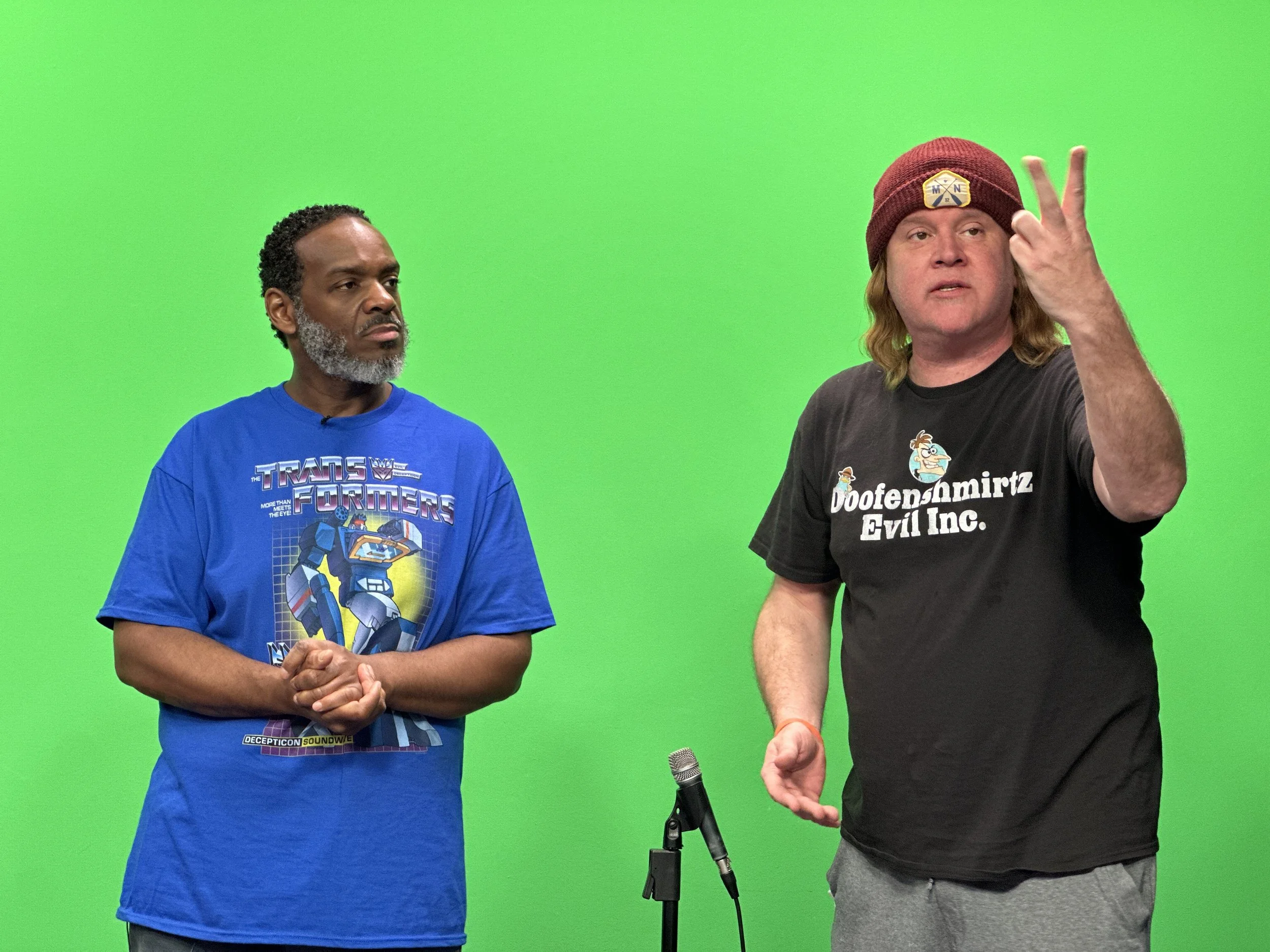 Two men standing in front of a green screen. The man on the left is wearing a blue T-shirt with a Transformers graphic and has short, curly hair and a beard. The man on the right is wearing a black T-shirt with a cartoon logo and text, a maroon beani