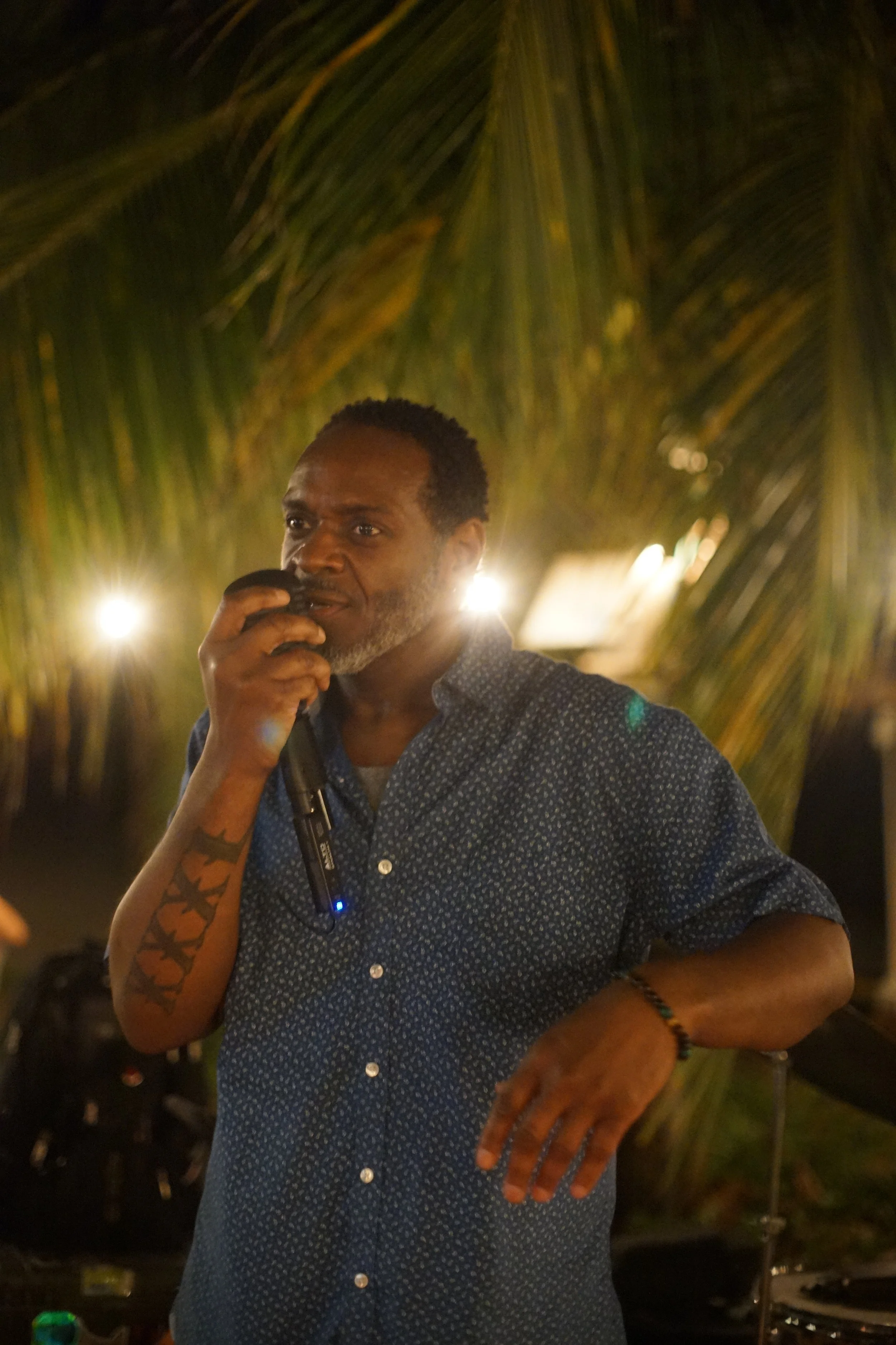 A man with dark skin and short hair, wearing a blue patterned shirt, holding a microphone, standing outdoors at night under string lights and palm trees.