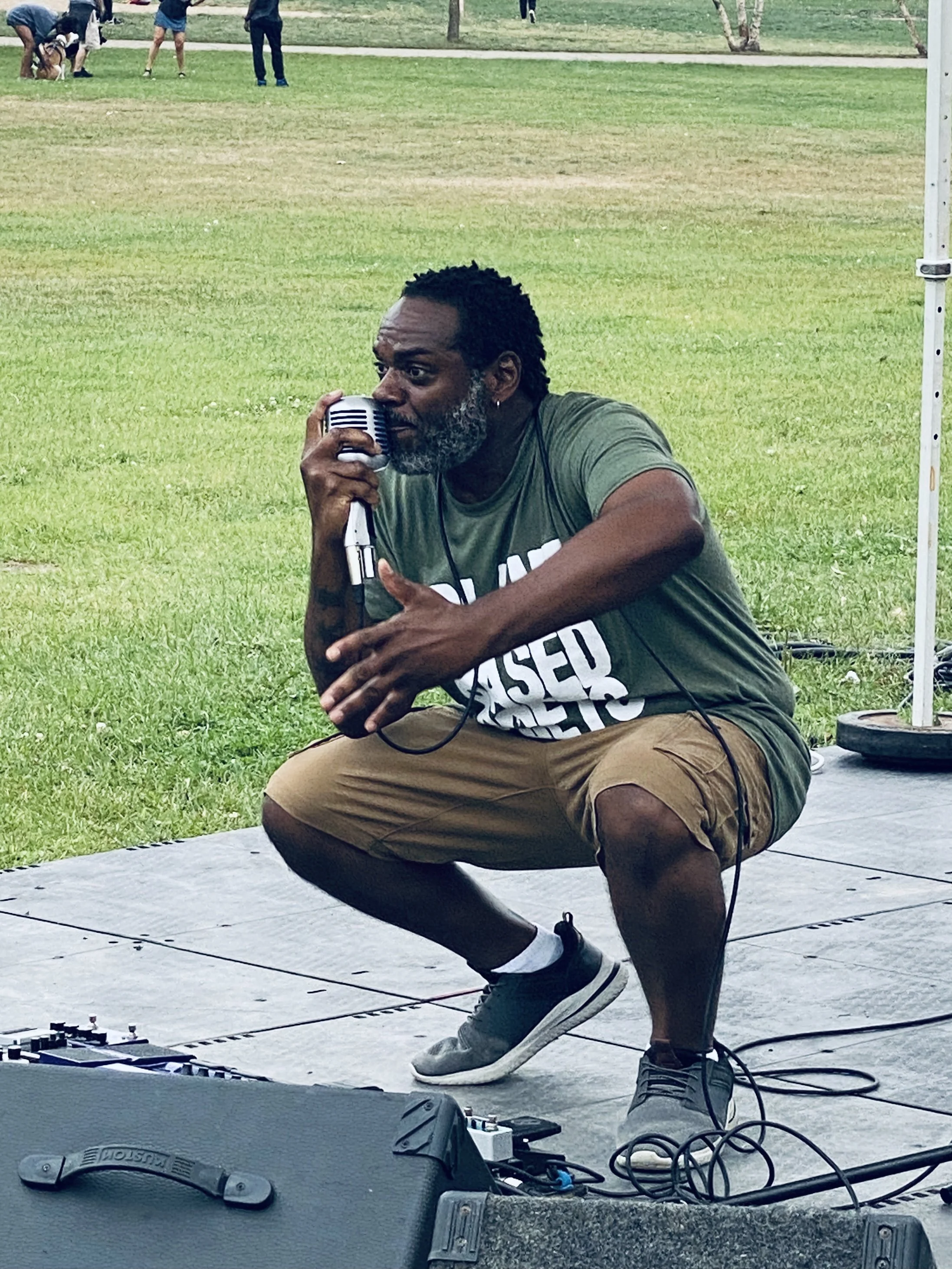 A man crouches on outdoor stage in front of music equipment, holding a microphone near his mouth, with musical gear and cables on the ground, and a grassy park with people in the background.