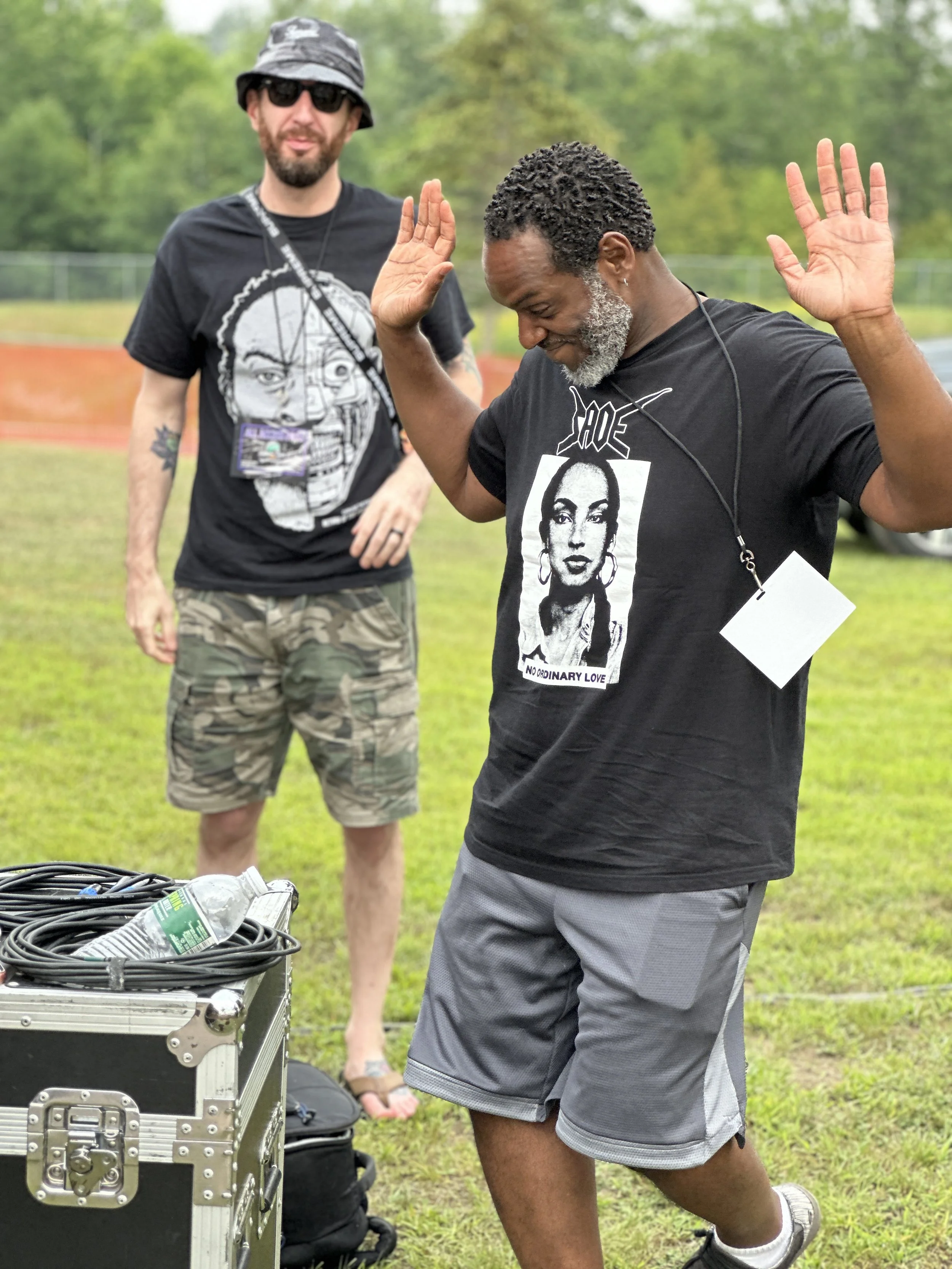 Two men outdoors, one with a beard, wearing sunglasses, a camouflage hat, and a T-shirt with a face graphic, stands in the background. The other man has gray hair and beard, wears a black T-shirt with artwork and the words "NO ORDINARY LOVE," gray sh
