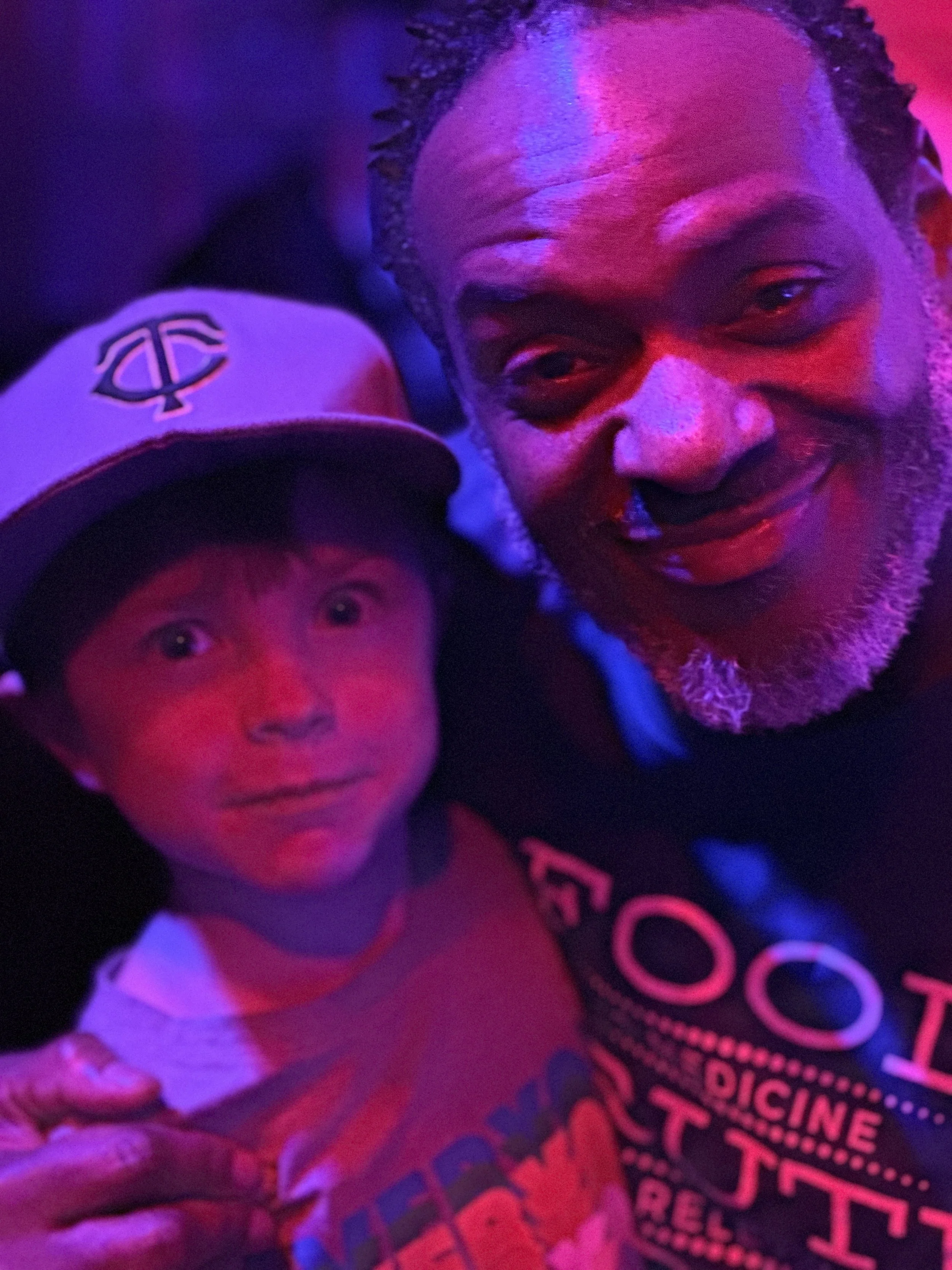 A young boy and an adult man are taking a selfie under colorful party lights, both smiling and looking at the camera. The boy is wearing a black hat and a white shirt with red and blue accents. The man has a beard and short hair.