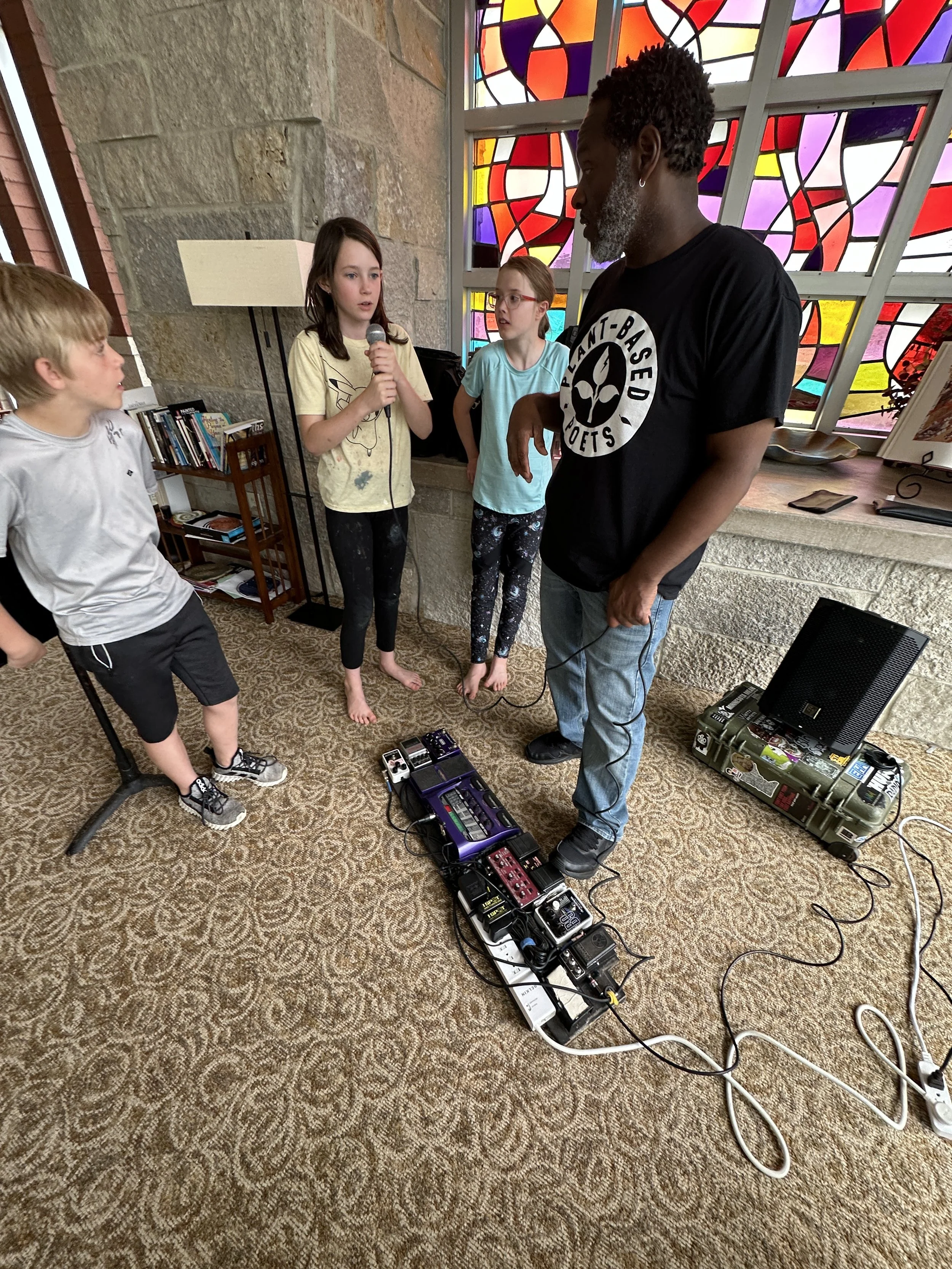 An adult man with gray hair and beard, wearing a black T-shirt with a 'Plant-Based Poets' logo, standing in front of three children, one of whom is speaking into a microphone. The children are barefoot, surrounded by musical equipment, in front of a 