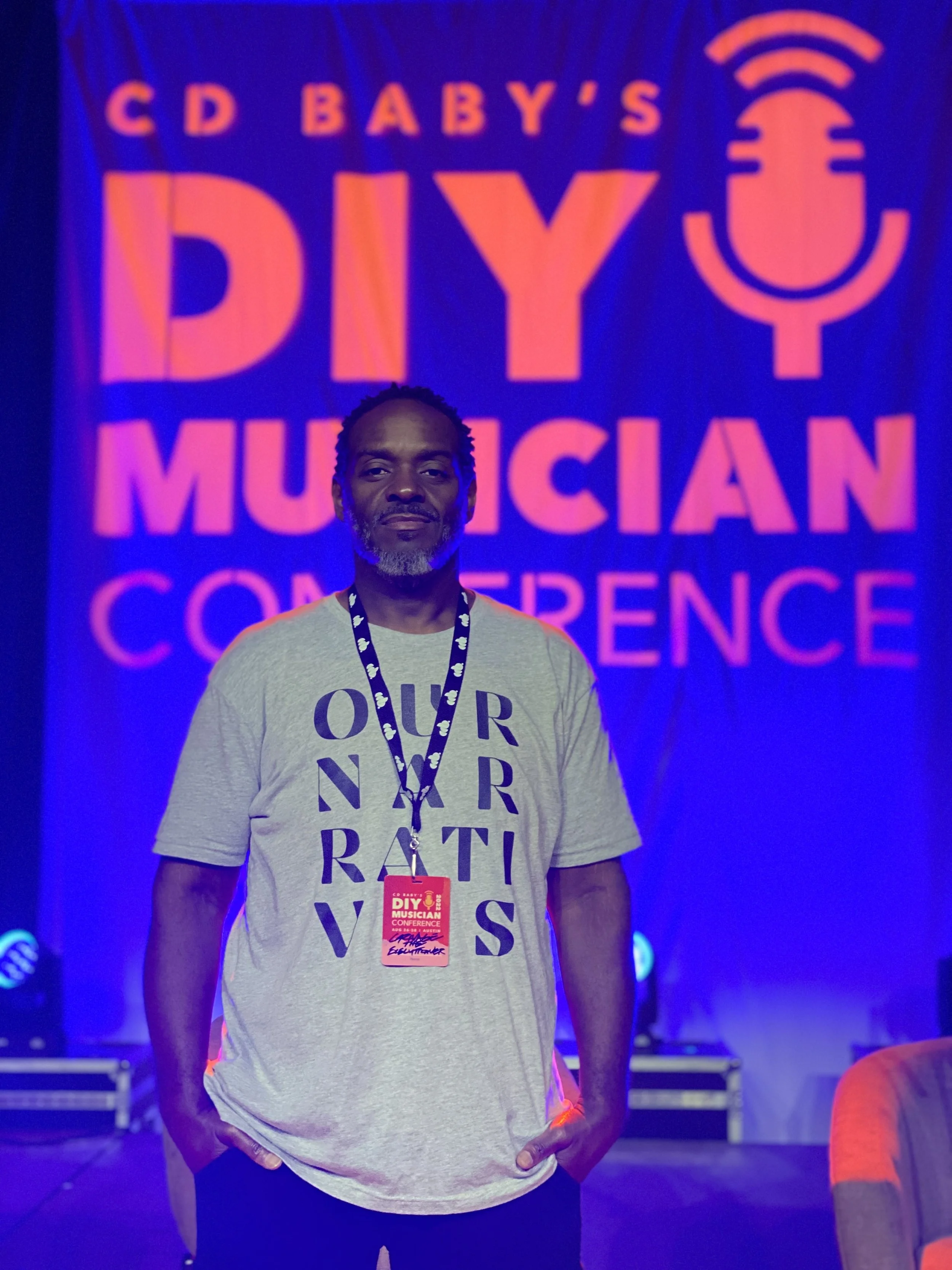 A man wearing a gray T-shirt with text, standing in front of a large, illuminated sign that reads 'CD Baby's DIY Musician Conference' with a microphone graphic. He has a conference badge hanging around his neck.