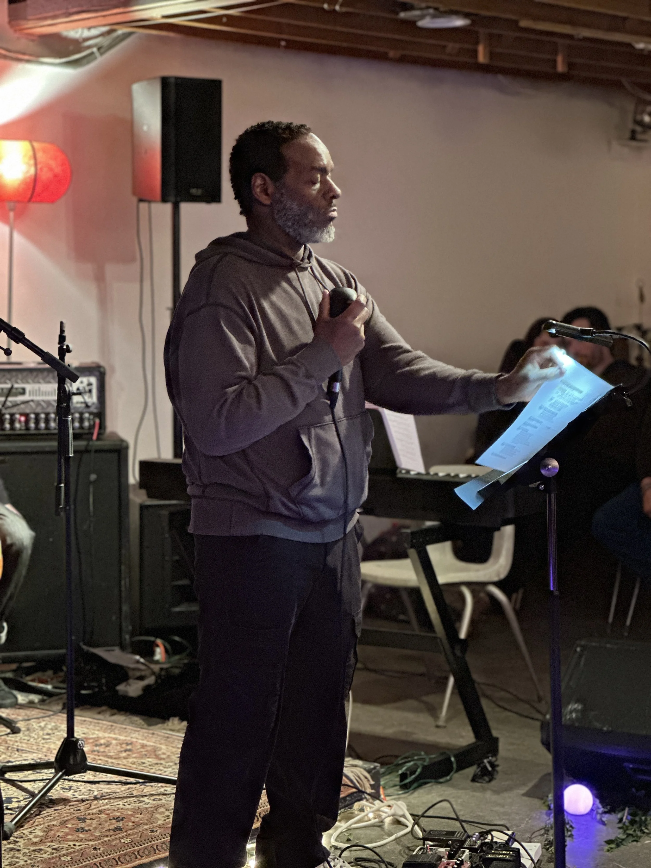 A man with a beard, wearing a hoodie, standing in a music or performance space with a tablet or sheet of music and a microphone in hand, surrounded by musical equipment and a speaker.
