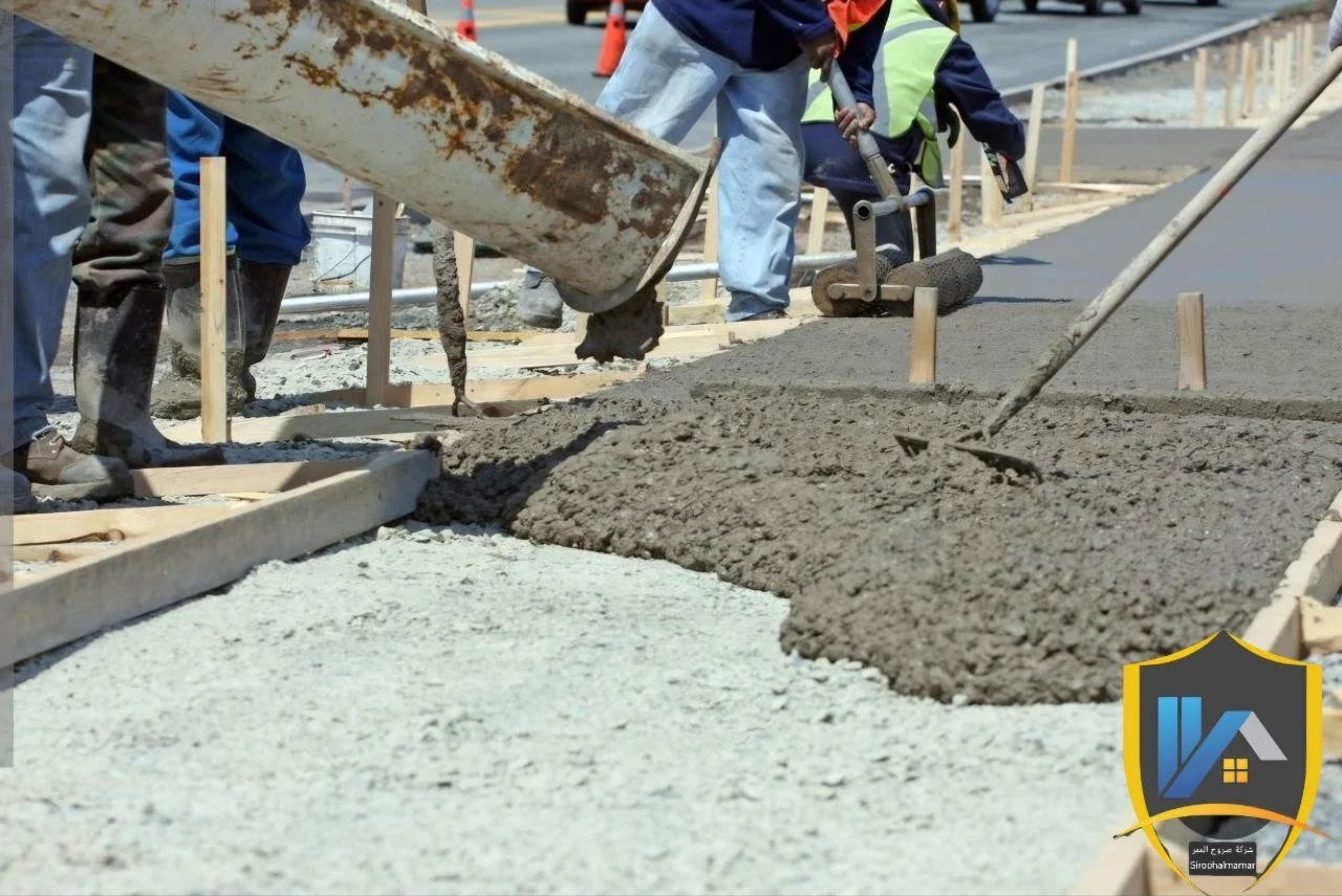 Workers pouring concrete on a sidewalk under construction, with wooden formwork and construction tools visible.