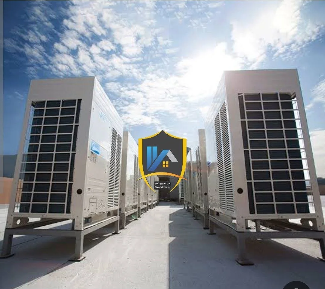Multiple large industrial air conditioning units on a rooftop under a partly cloudy sky. A logo with a house and shield is centered.