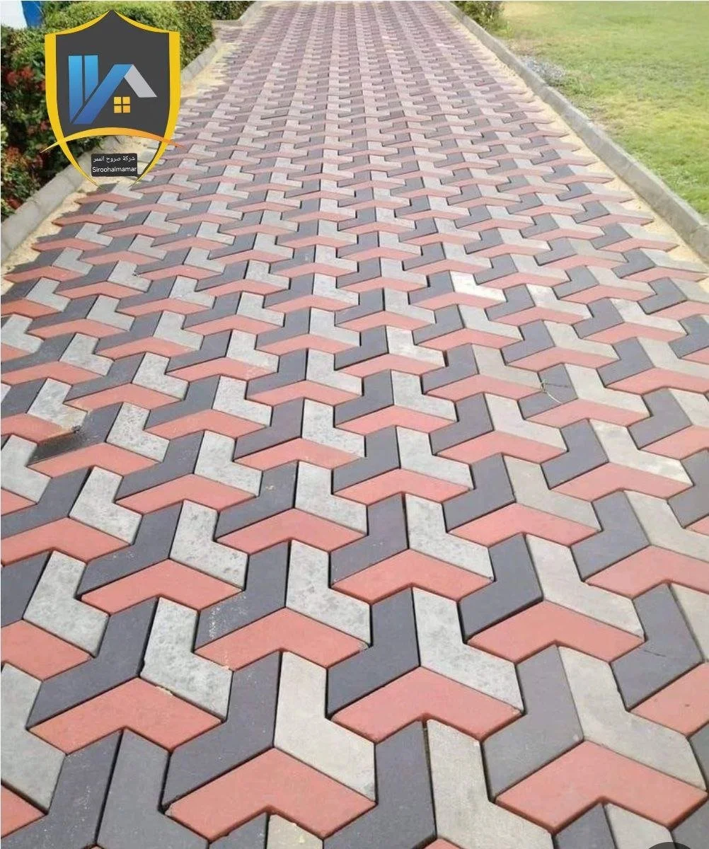 A paved walkway with a repeating geometric pattern of interlocking hexagonal tiles in red, black, and gray colors, bordered by grass and small plants.