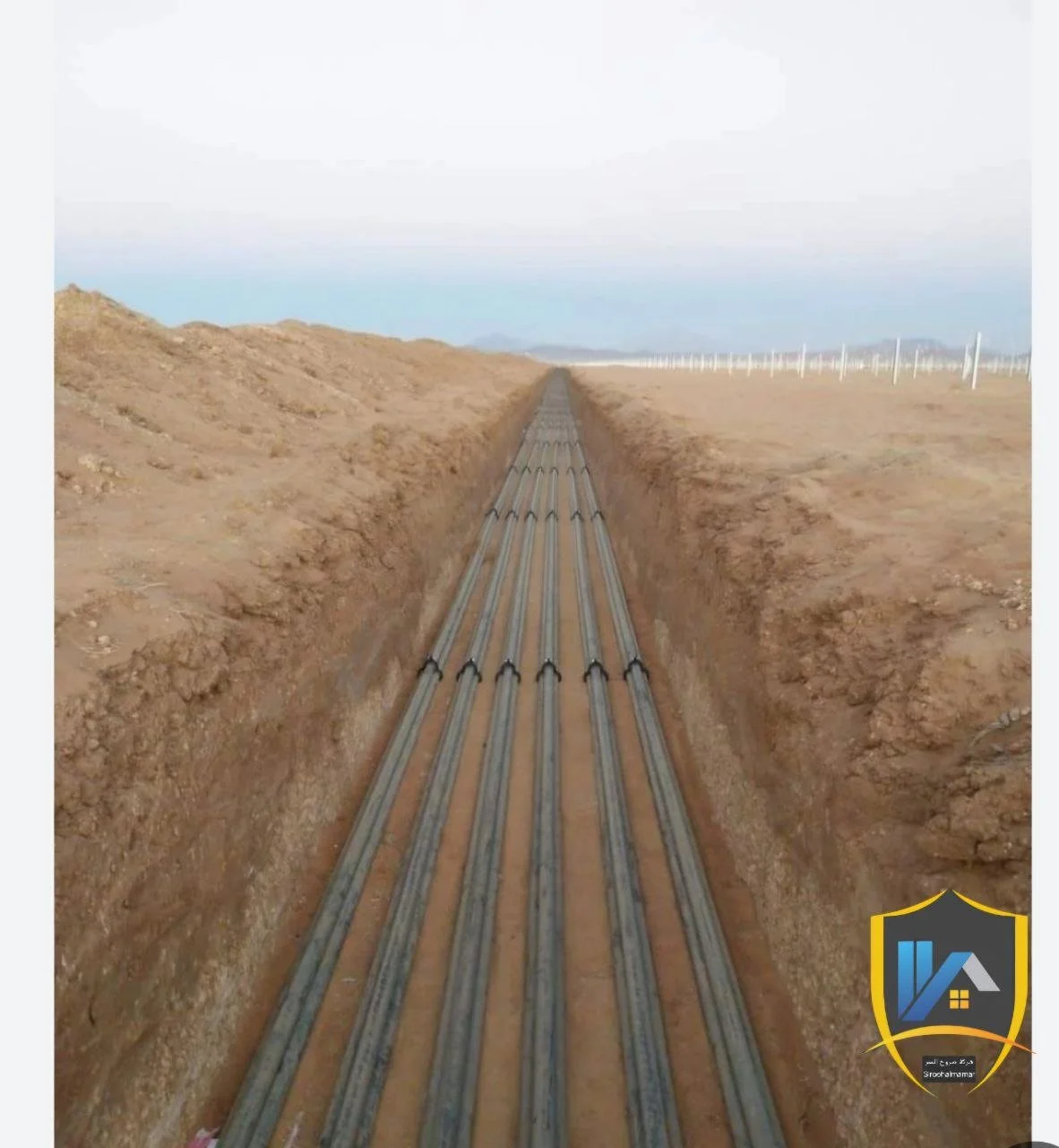 Multiple black pipes installed in a trench through a desert landscape, with wind turbines visible in the distance under a cloudy sky.