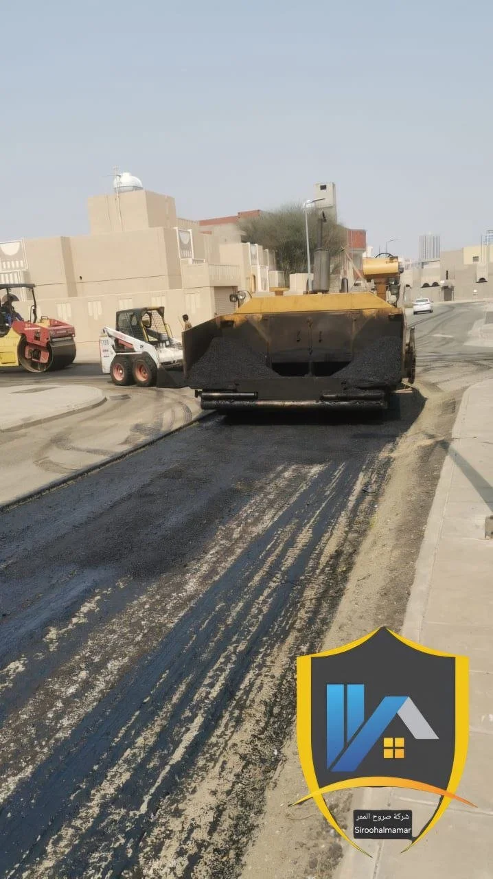 Asphalt paving process with a large road roller compacting the new road surface, alongside other construction machinery on an urban street with residential buildings and clear sky.