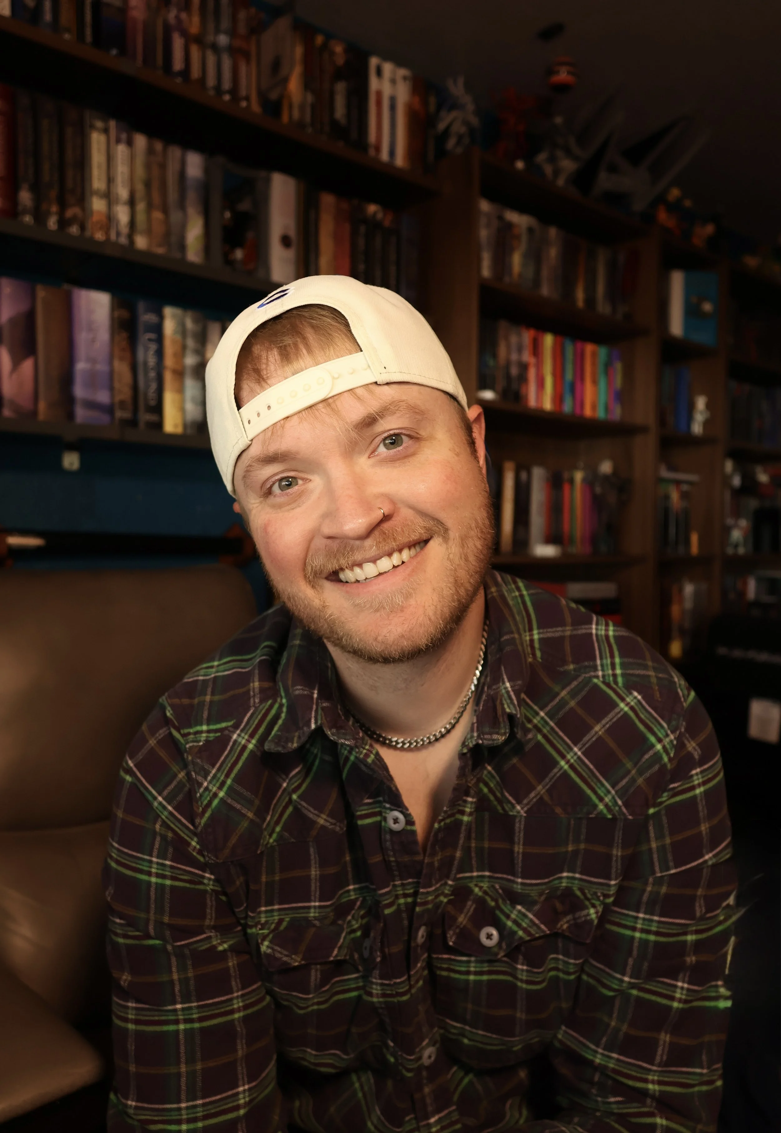 A smiling young man with a nose piercing wearing a white baseball cap backward and a checkered shirt, sitting in a room with bookshelves filled with books in the background.