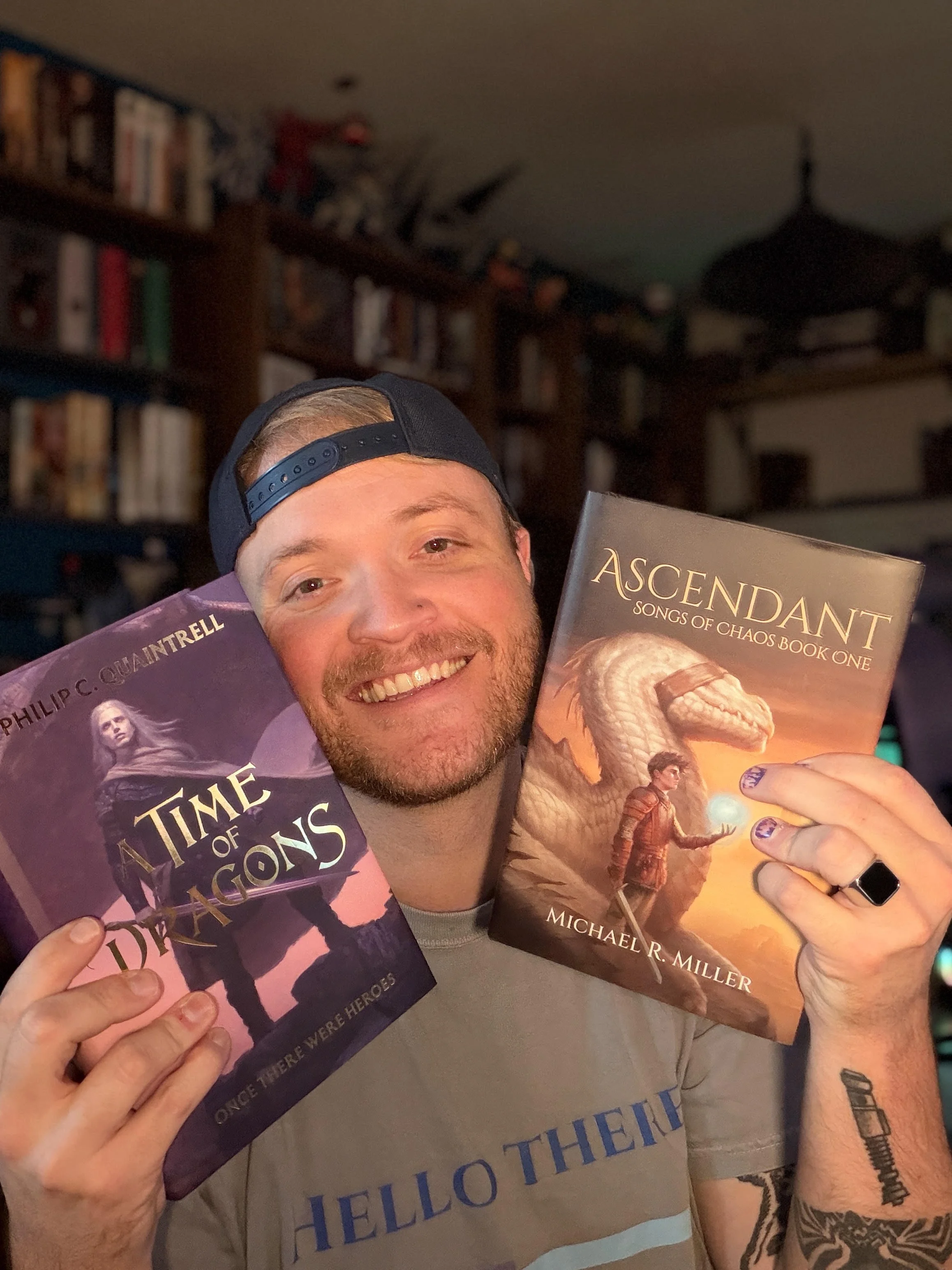A man smiling and holding two books titled 'The Time of Dragons' and 'Ascendant: Songs of Chaos Book One' in a room with a bookshelf and a ceiling fan in the background.