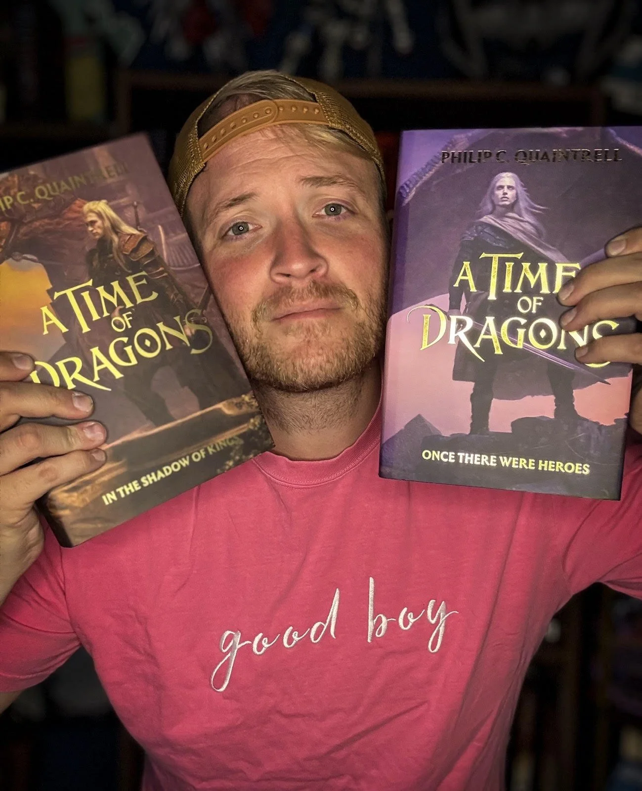 A man wearing a pink shirt and a gold cap is holding two copies of the book "A Time of Dragons" by Philip C. Quatrrell, one in each hand, with a background of shelves.