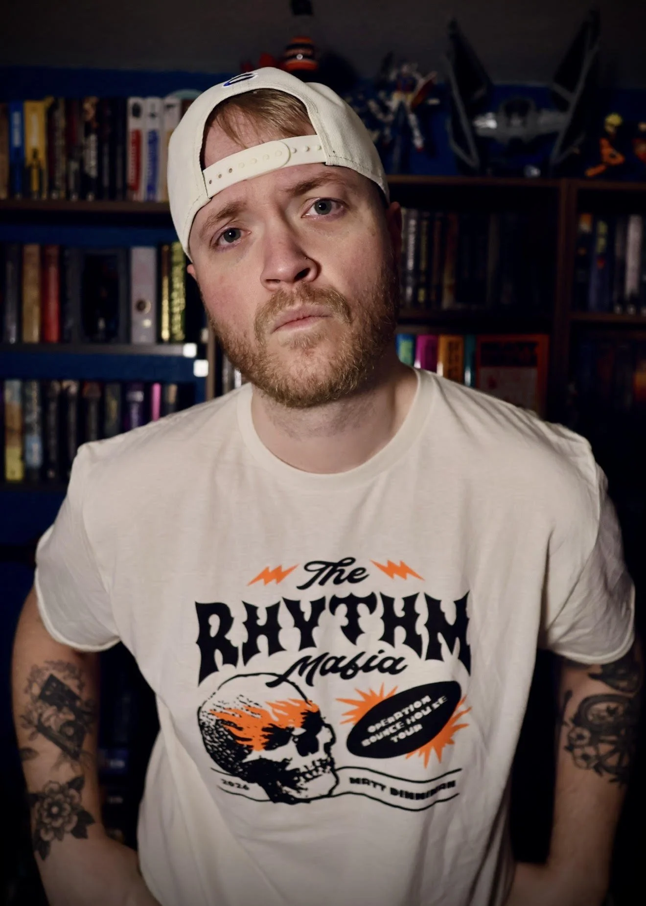 A young man with a beard and tattoos wearing a white t-shirt and a backwards baseball cap, sitting in front of a bookshelf with video games and models.
