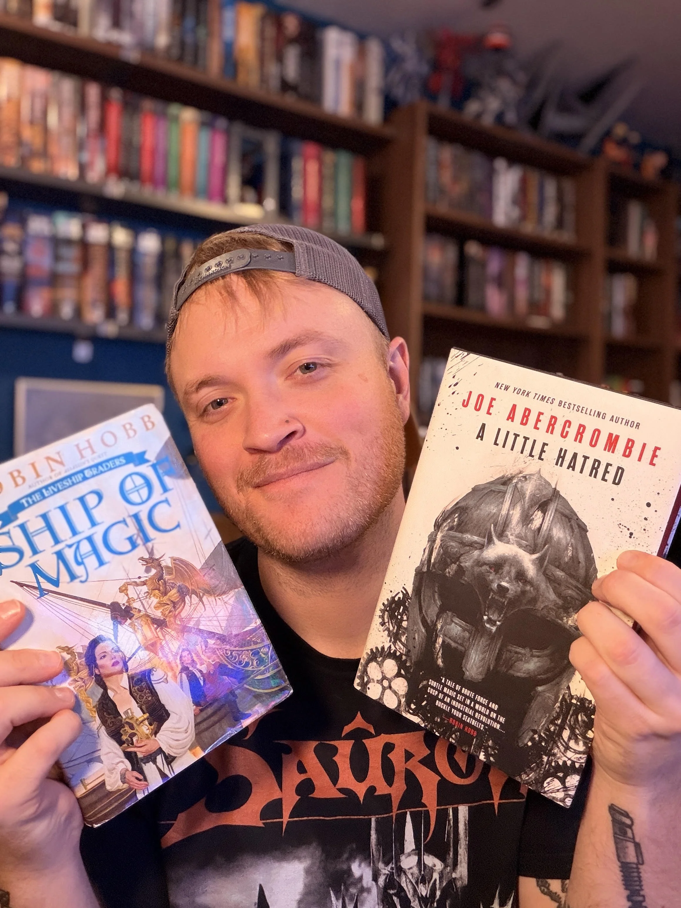 A young man wearing a backwards cap is holding two books in front of a background of bookshelves filled with various books. His face is partially visible, smiling slightly, and he has tattoos on his arms.