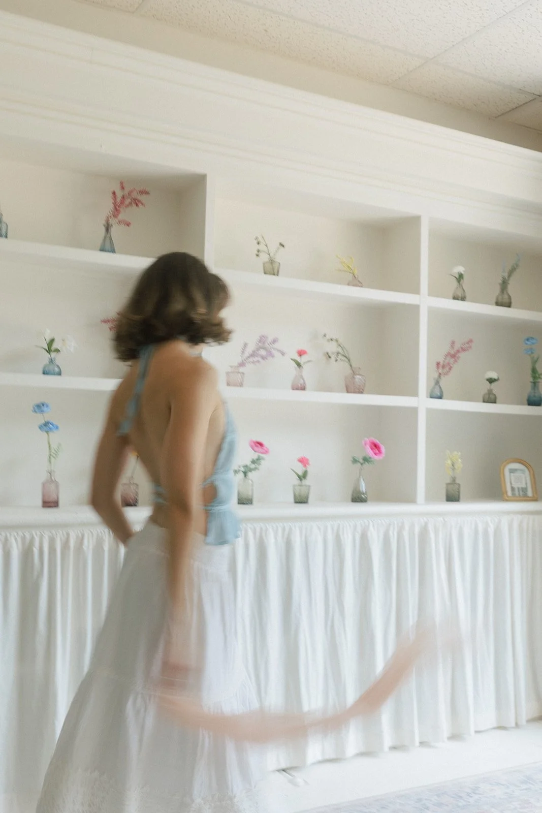 A woman with brown hair in a light blue top and white skirt standing in front of a white shelf with flower vases.