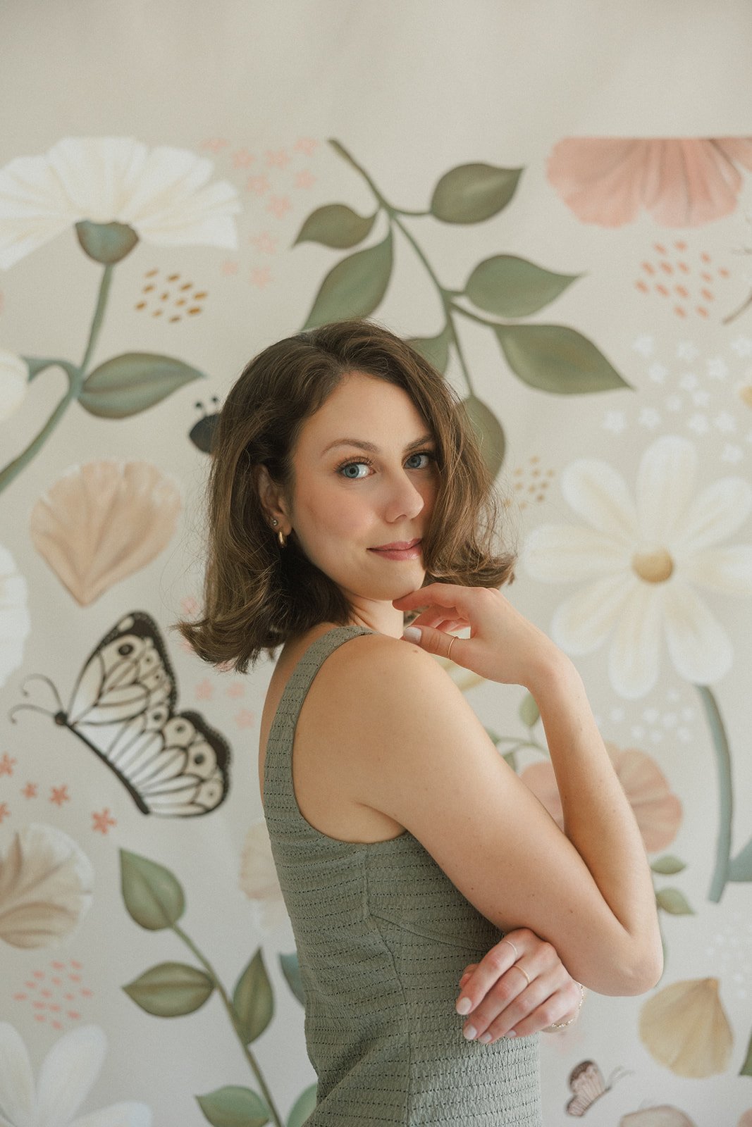 A woman with shoulder-length brown hair and blue eyes, wearing a sleeveless light gray dress, standing in front of a floral wallpaper with large flowers and butterflies, posing with a slight smile and hand near her chin.