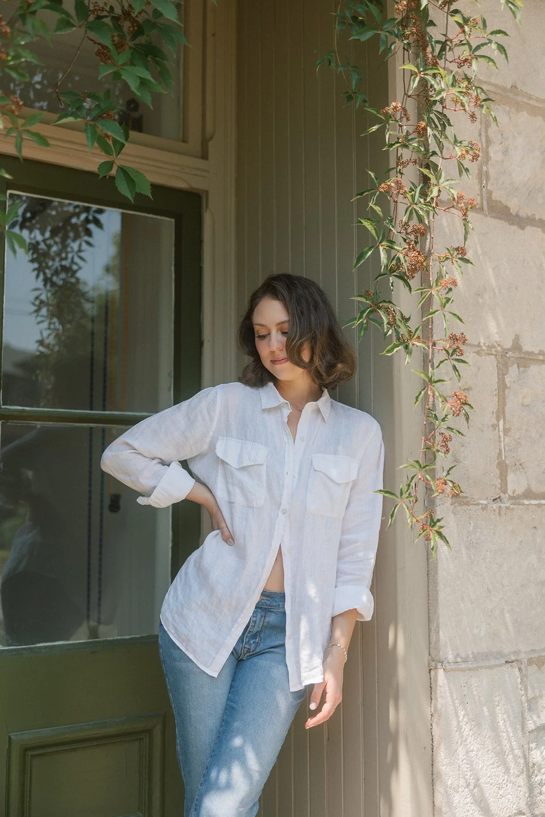 A woman standing outside leaning against a stone wall next to a green door with a window. She is wearing a white button-up shirt and blue jeans, with her right hand on her waist. Green vine plants with small leaves and reddish flowers grow along the wall and frame part of the scene.