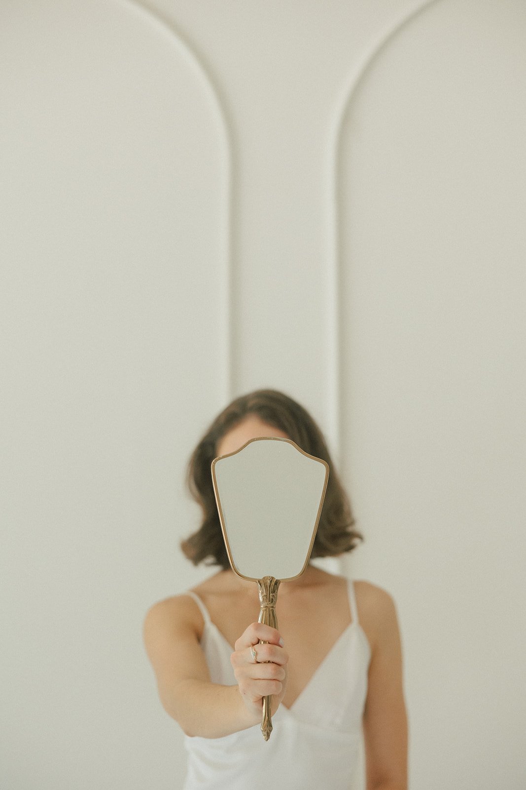 A woman with shoulder-length dark hair holding a small, ornate mirror in front of her face, wearing a white sleeveless top, standing in front of a mirror with a light-colored wall background.