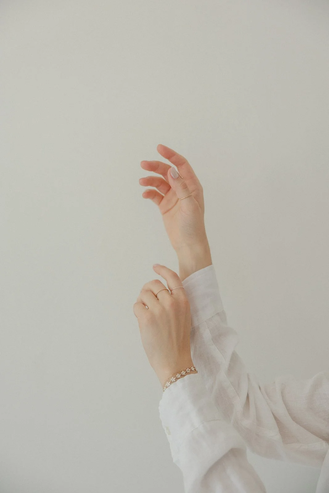 Two hands with rings and a bracelet, one slightly above the other, against a plain white wall, with light-colored long sleeves.