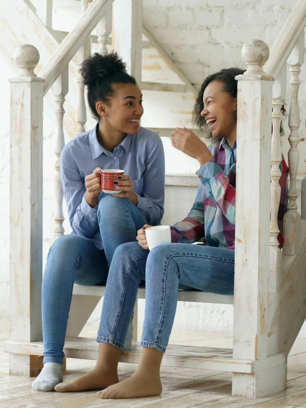 Two women sitting on a staircase, smiling and talking while holding mugs.