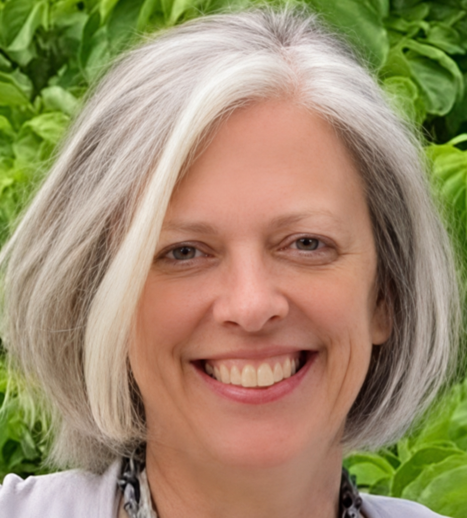 A woman with shoulder-length gray and white hair smiling, standing outdoors with green leafy plants in the background.