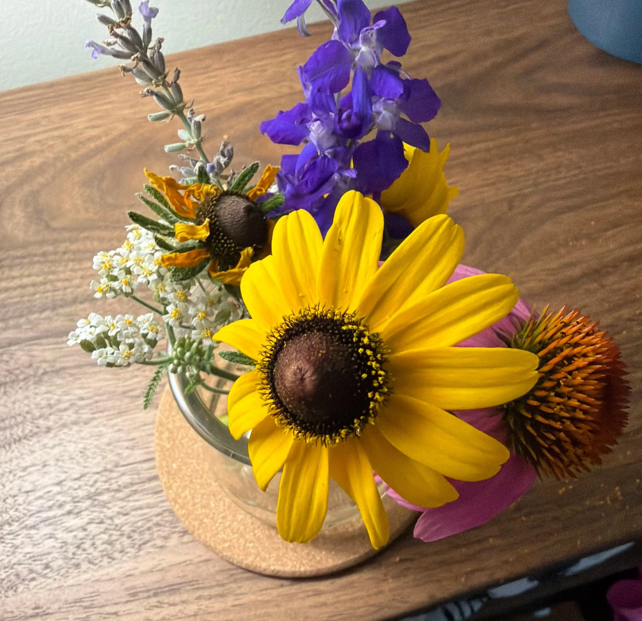 A bouquet of colorful flowers on a wooden table, featuring a large yellow sunflower, purple flowers, white flowers, and a bee collecting nectar.