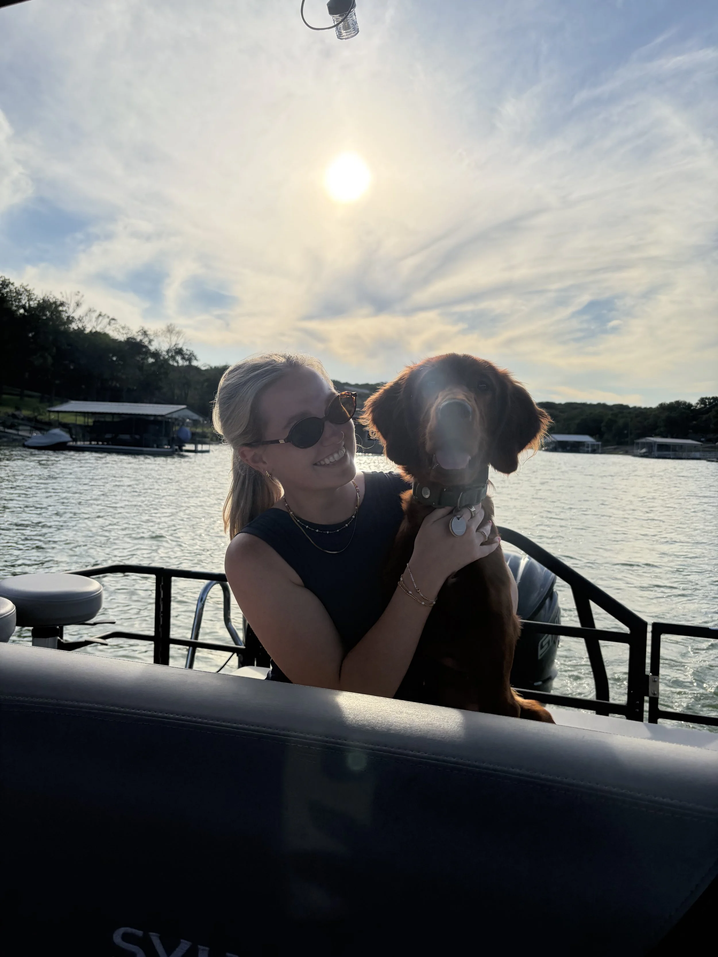 A woman with blonde hair, wearing sunglasses, holding a brown dog with a collar, on a boat on a lake during sunset with a cloudy sky and trees in the background.