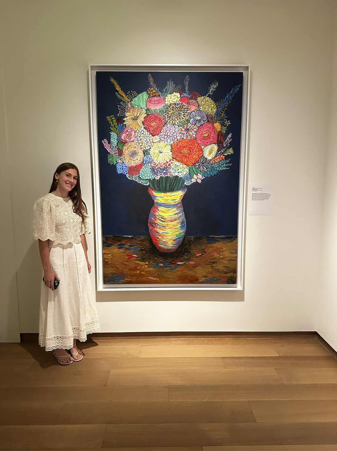 A woman in a white dress standing next to a large colorful painting of a flower bouquet in a vase at an art gallery.