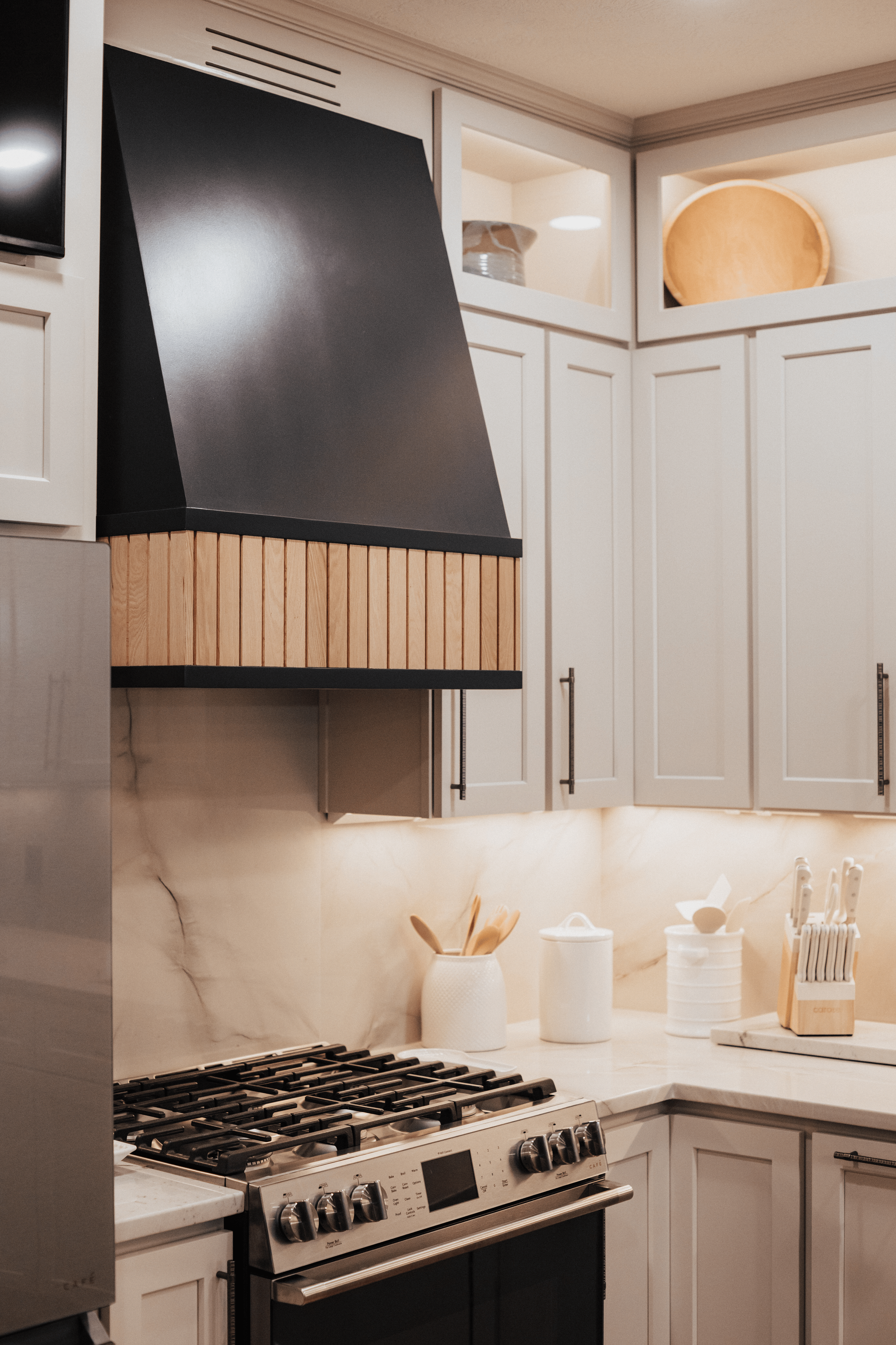 modern kitchen with custom matte black range hood with wood slat band surrounded by light grey cabinets and white marble patterned tile backsplash