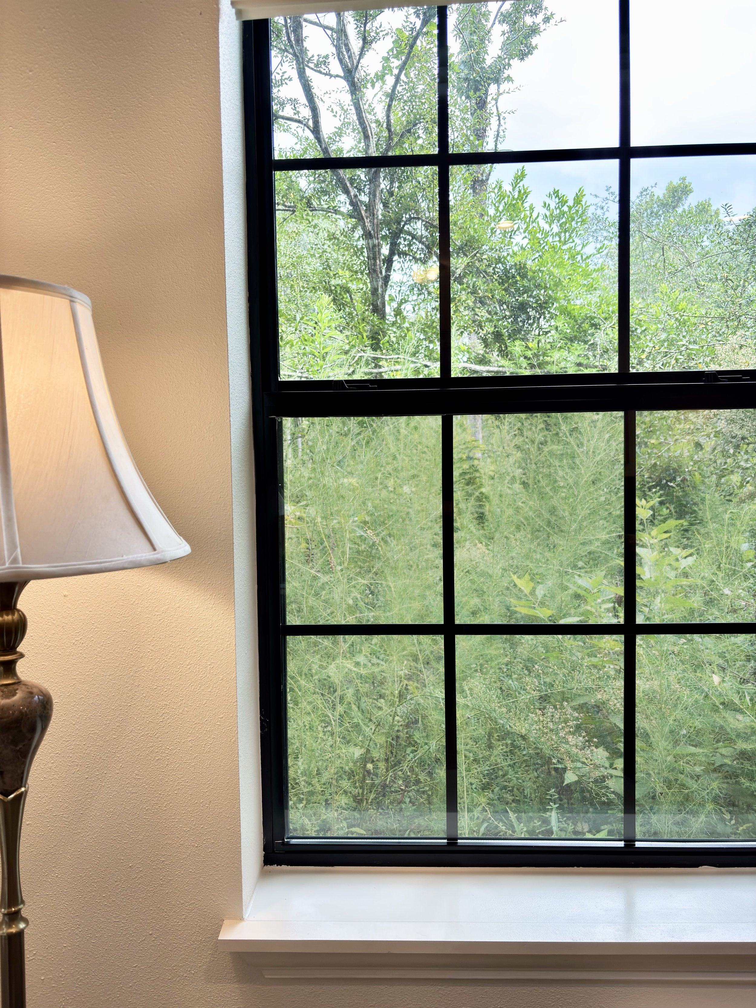 A window with black grid panes looking out onto green trees and foliage outside. Part of a beige wall and a white window sill are visible, along with a brass and cream-colored lamp on the left.
