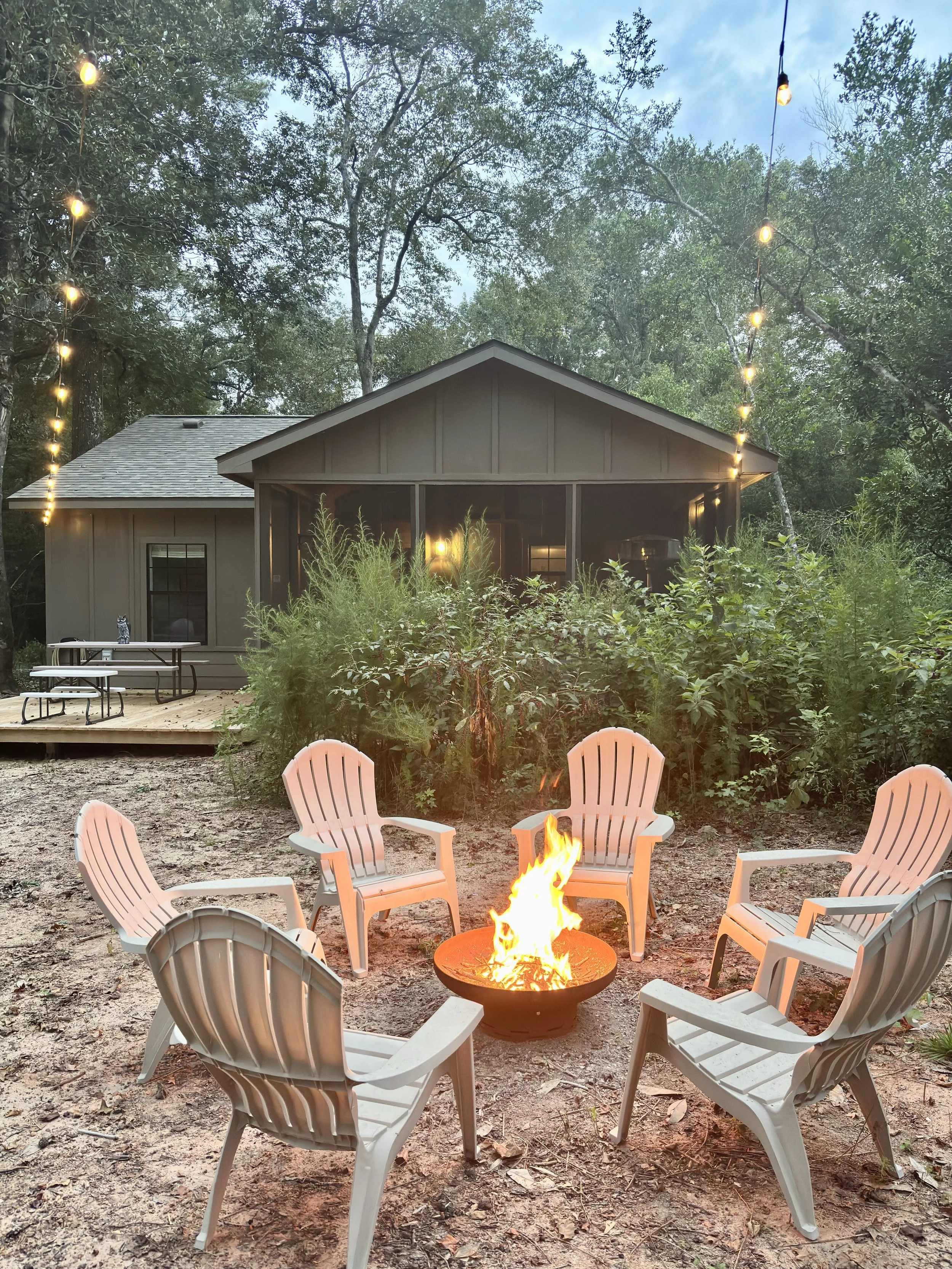 A backyard scene with a fire pit surrounded by taupe plastic Adirondack chairs, a house in the background, and string lights hanging from trees, during early evening or dusk.