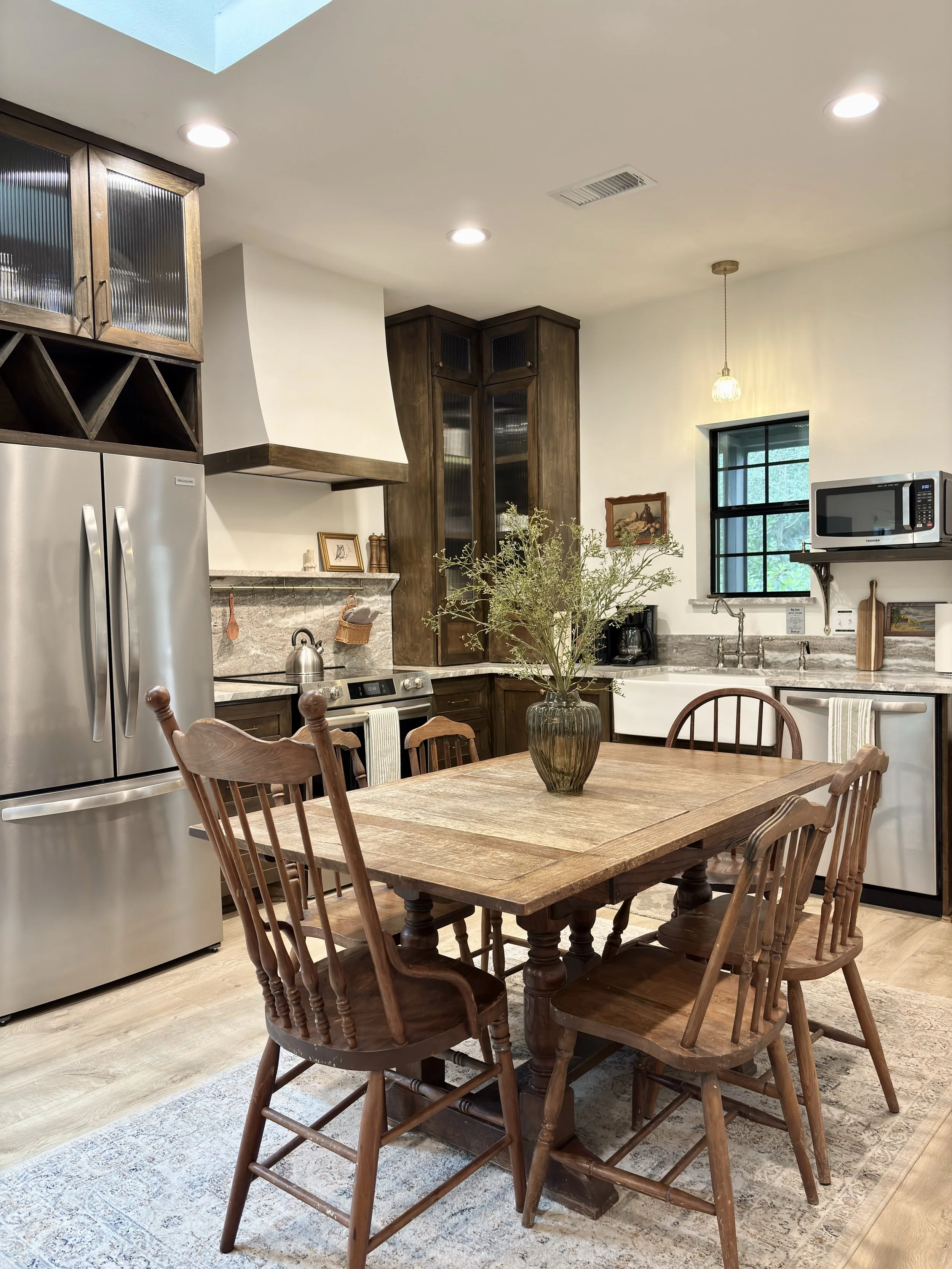 A wooden dining table with six matching wooden chairs in a kitchen, a vase with greenery in the center, stainless steel appliances, dark wooden cabinetry, a window, and a small pendant light.