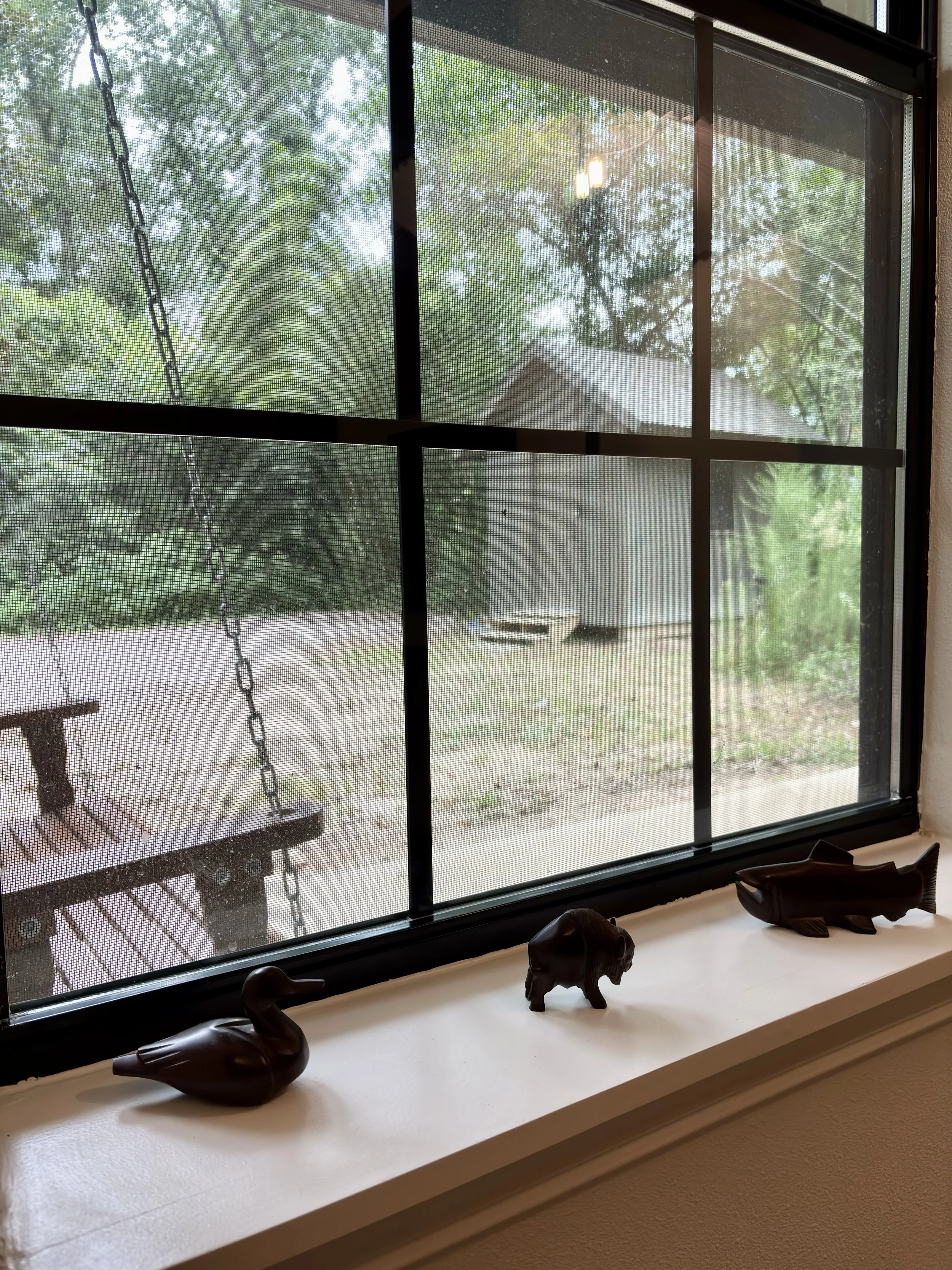 View of a backyard through a window with a screen, showing trees, a house, and a wooden deck outside, as well as decorative animal figurines on the windowsill inside.