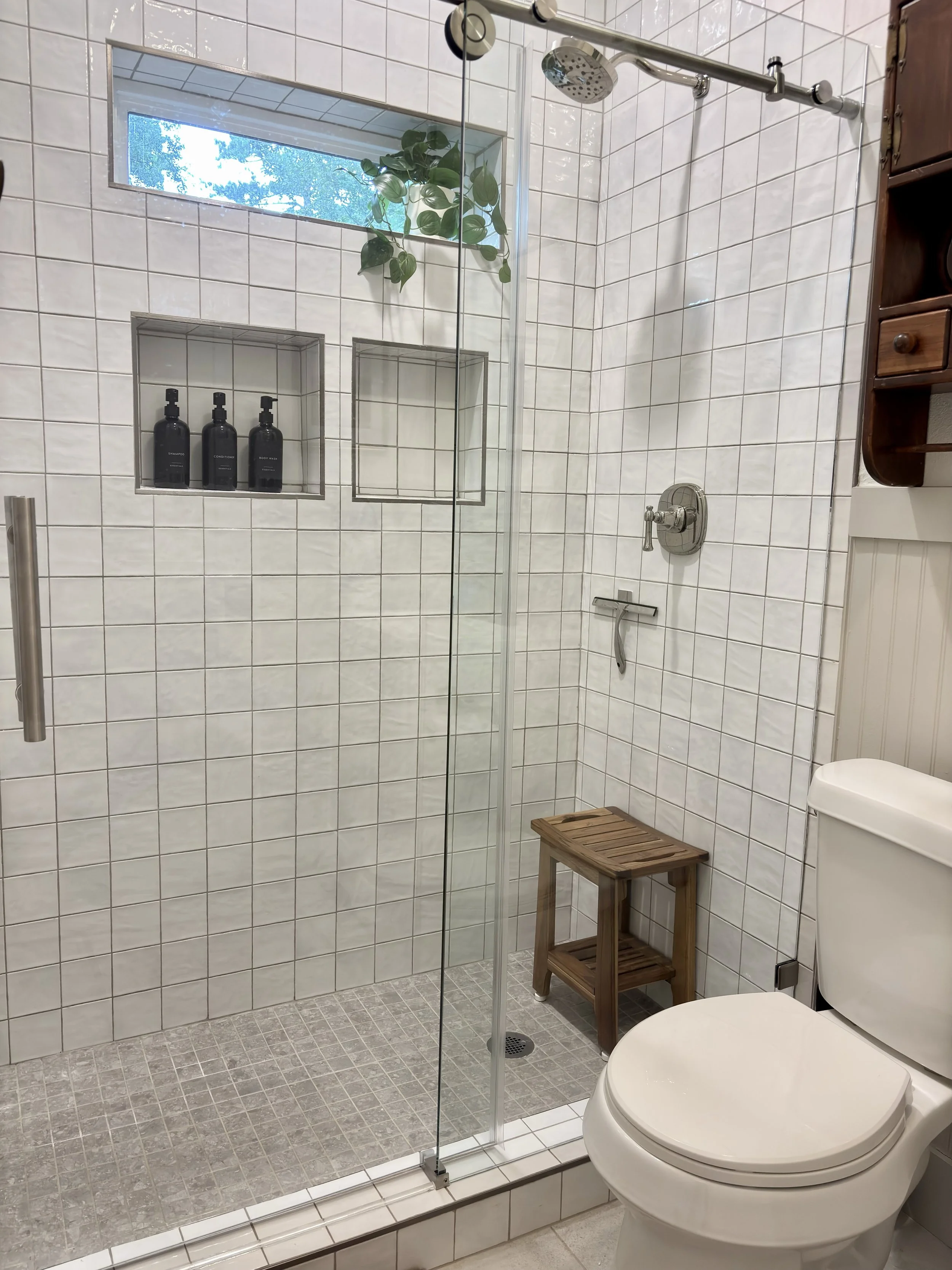 A modern bathroom with a glass-enclosed shower area, white tiled walls, a potted plant, built-in shelves with toiletries, a small wooden stool, and a white toilet.