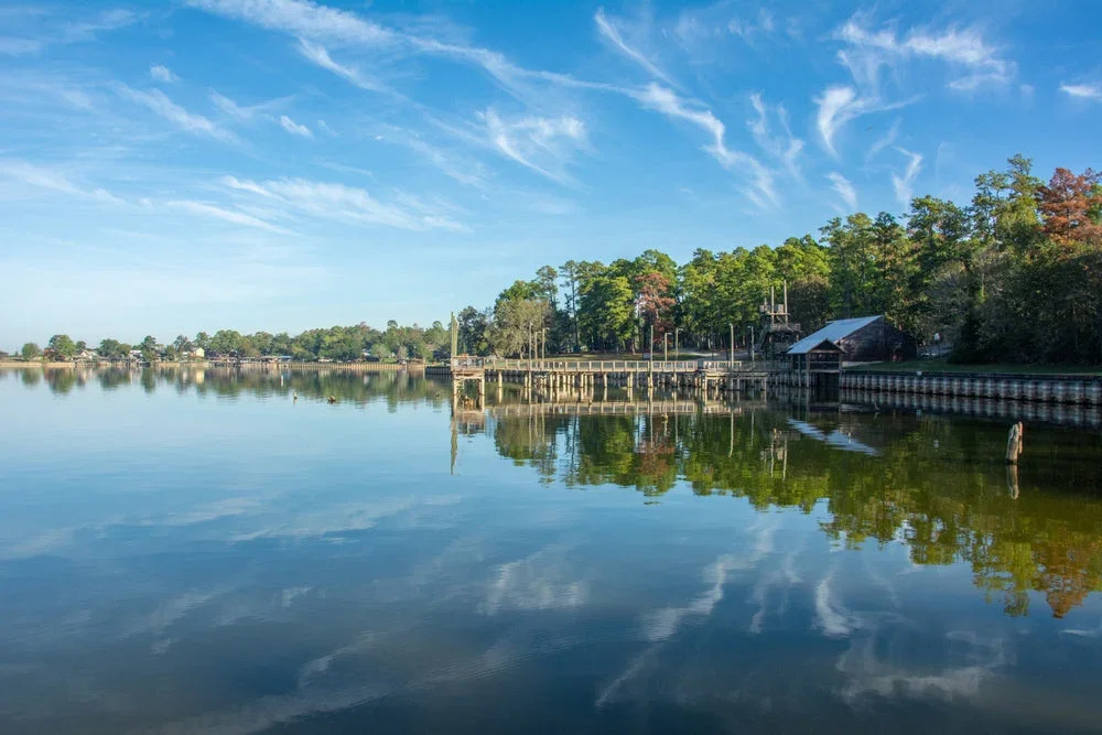 Calm river with reflections of trees and blue sky with wispy clouds, shoreline with wooden pier and small building.