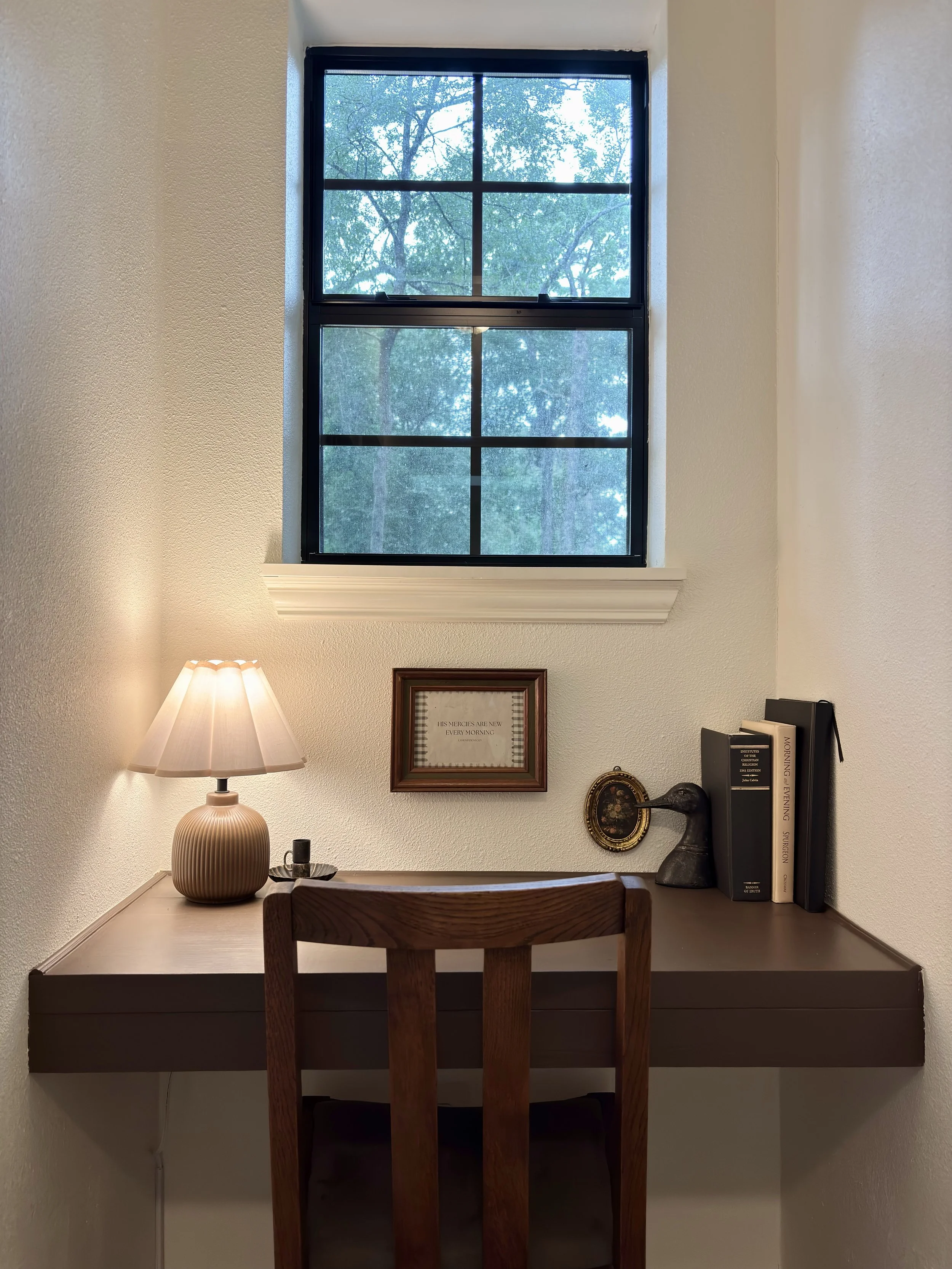A cozy corner with a wooden desk, a table lamp, framed quote, books, a decorative frame, and a small sculpture under a double-hung window showing green trees outside.