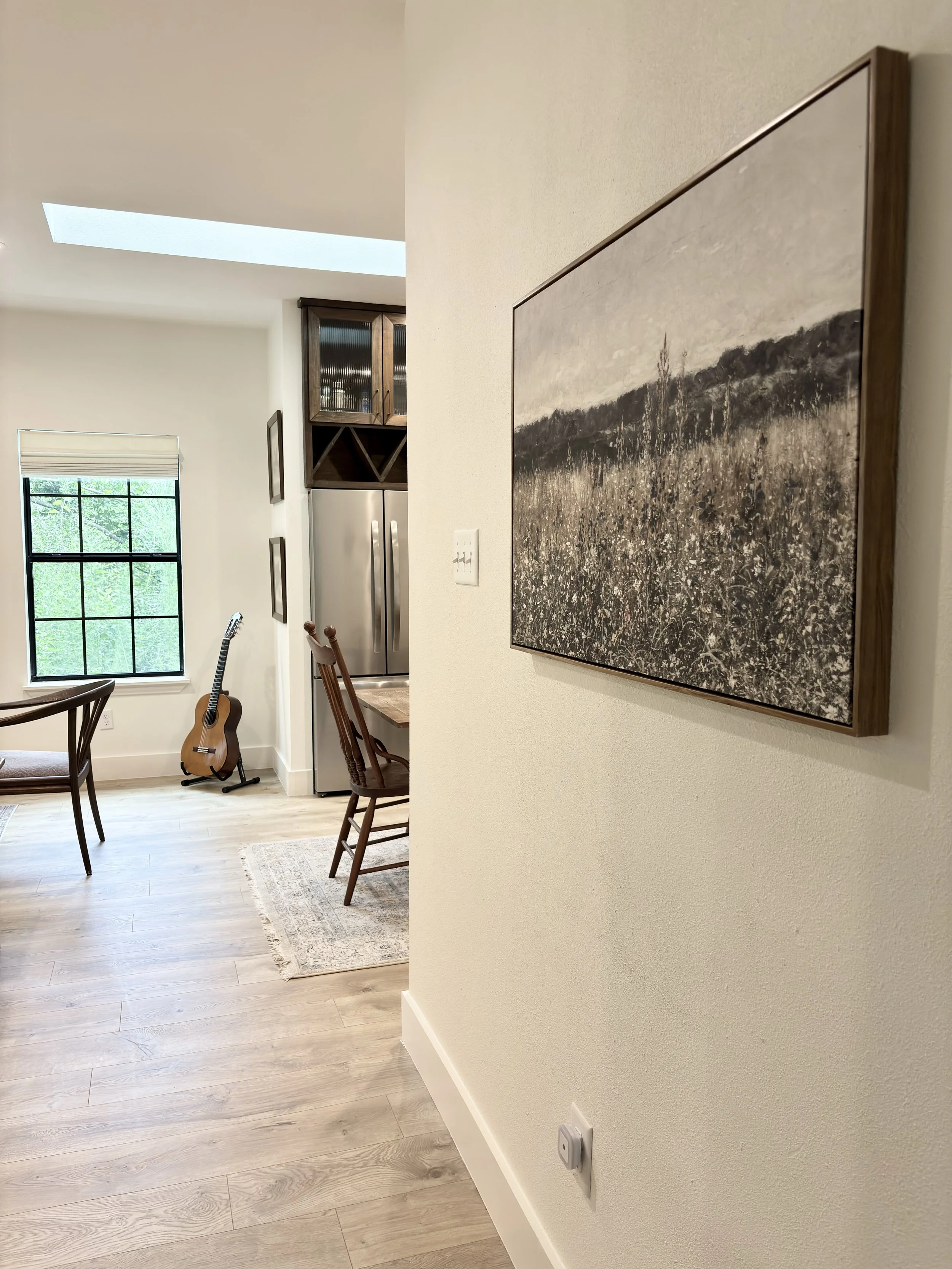 Interior view of a home with a hardwood floor, a painting on the wall, a window with greenery outside, a guitar leaning against the wall, a dining table with chairs, a kitchen with a refrigerator, and a skylight ceiling.