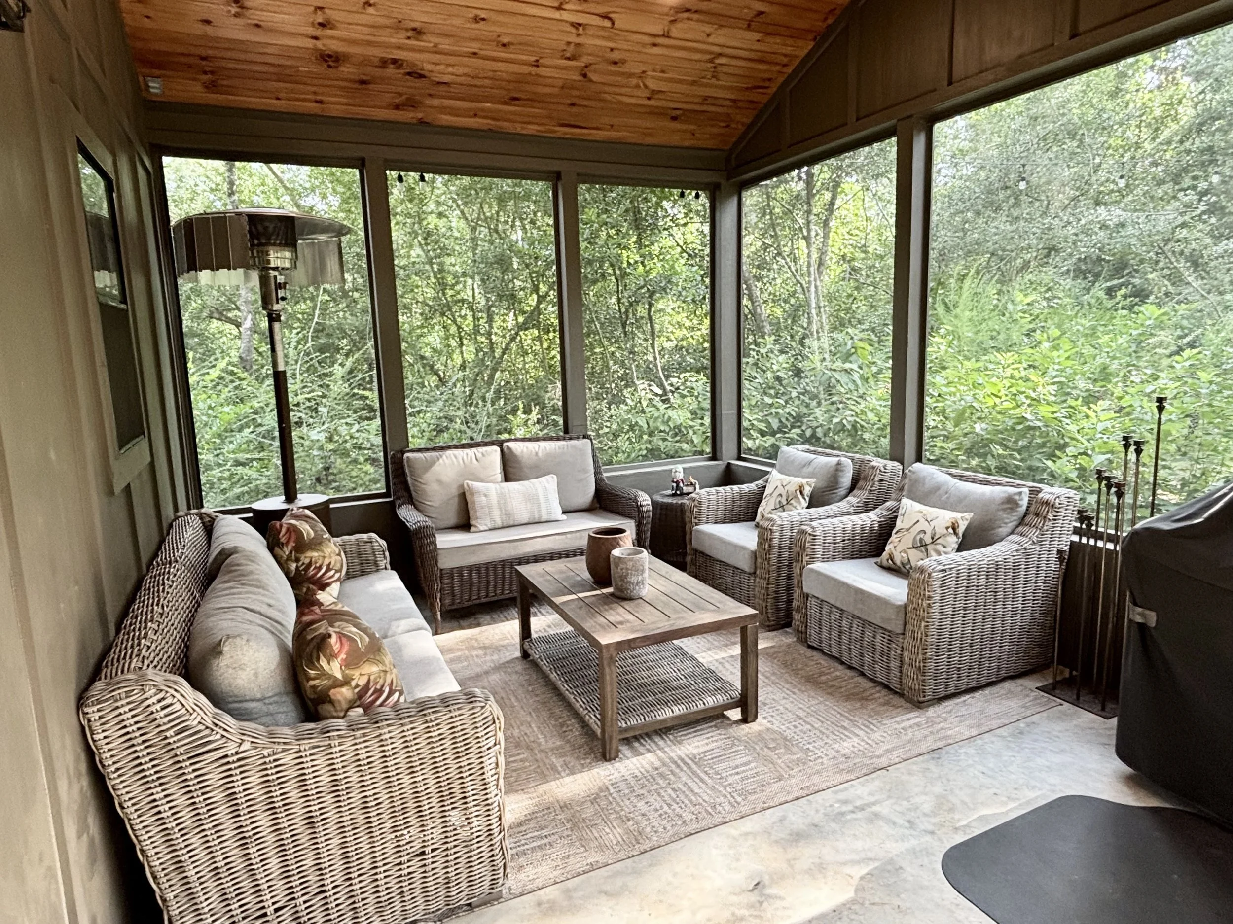 Sunroom with wicker and upholstered seating, a wooden coffee table, and view of greenery outside through large windows.