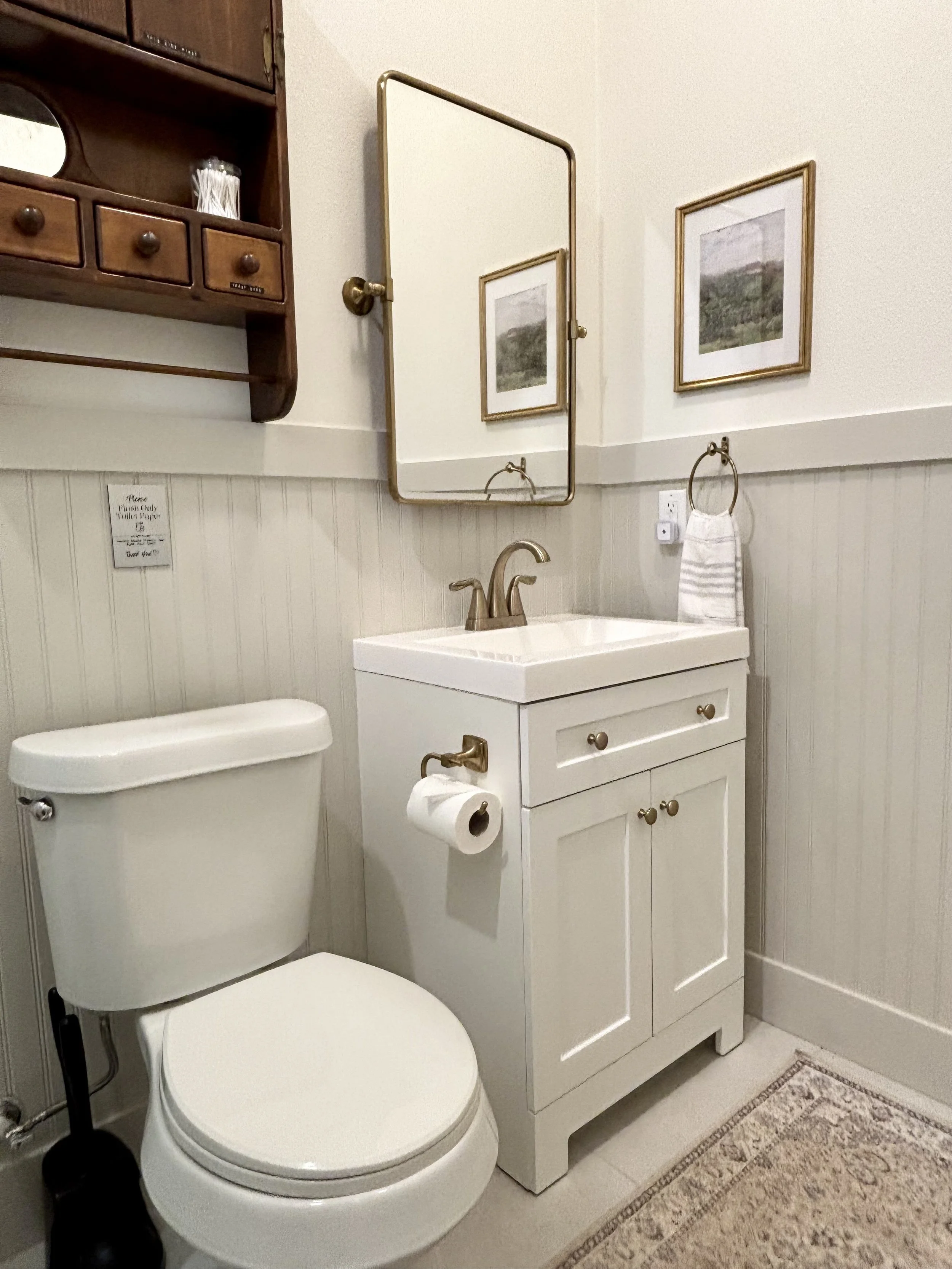 A bathroom featuring a white toilet, a small white vanity with a golden faucet, a mirror above the sink, framed landscape artwork on the wall, a wooden wall-mounted cabinet, a towel ring with a gray striped towel, a wall outlet, a toilet paper holder, and a beige rug.