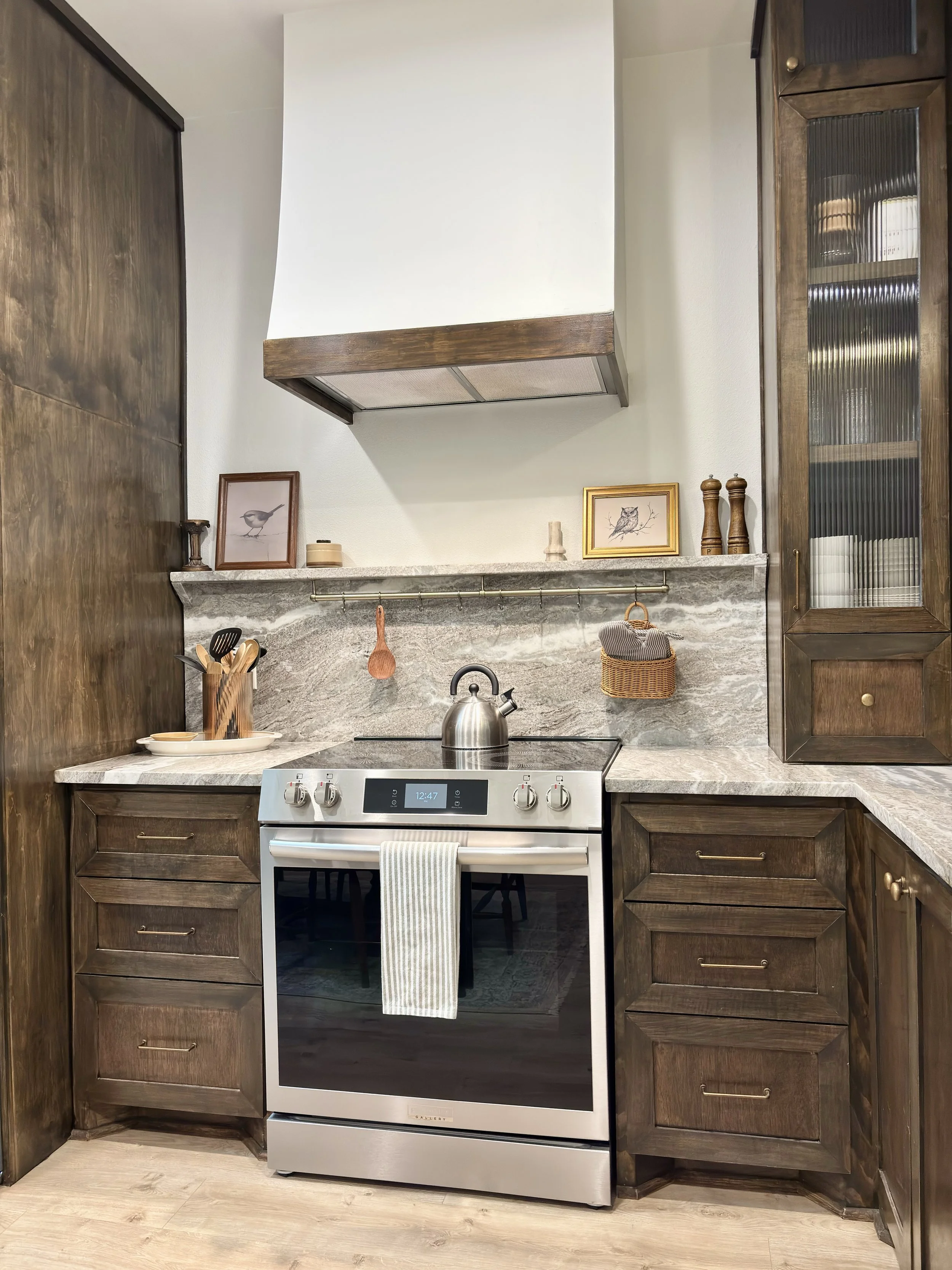 A kitchen with dark wooden cabinets, a marble countertop, and stainless steel stove with a digital clock reading 12:47. Decor includes framed artwork of a bird and an owl, wooden utensils in a holder, and a spice grinder. There's also a wicker basket and some small containers.