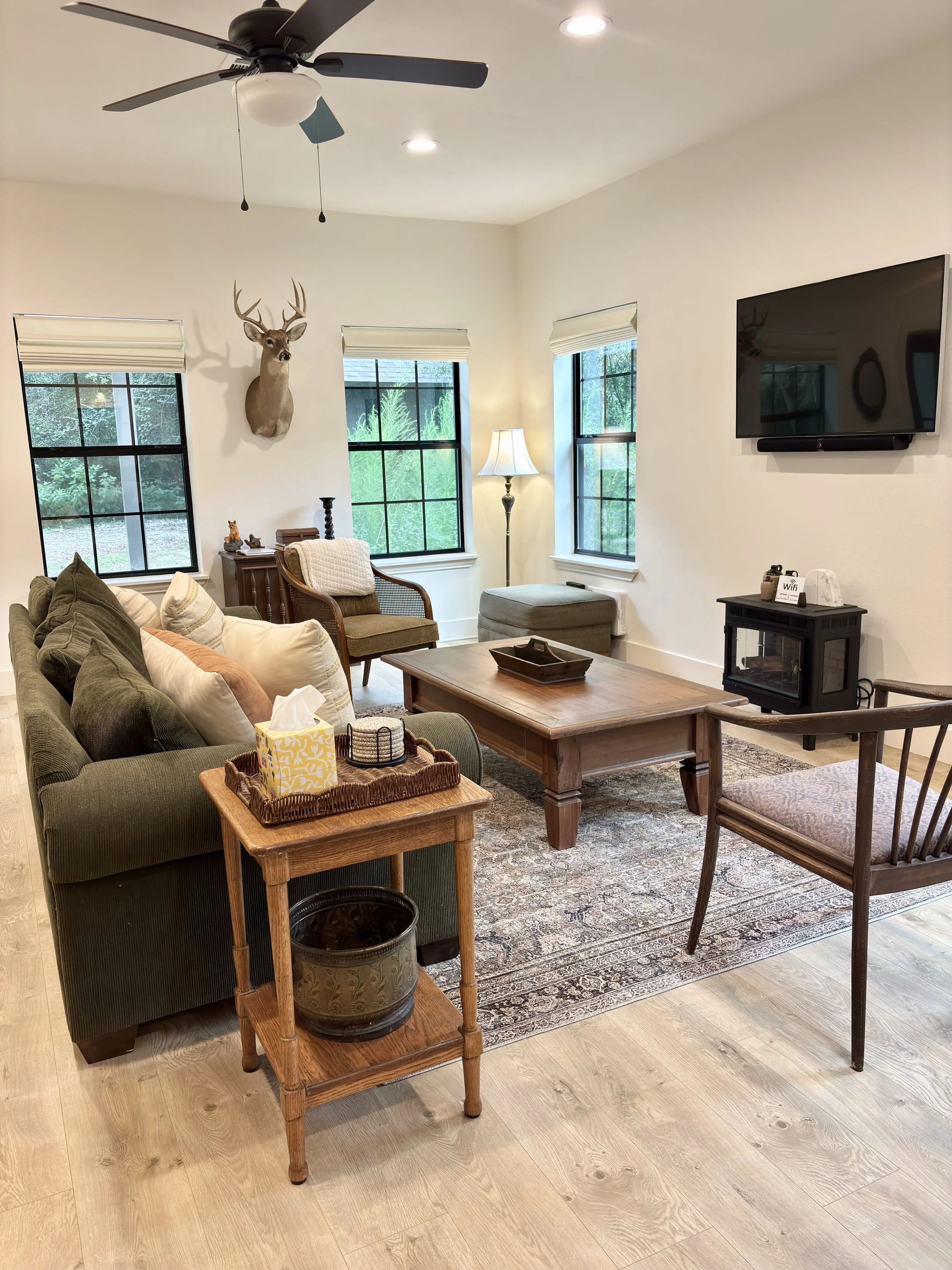 A living room with a green sofa, brown chairs, a wooden coffee table, a rug, a wall-mounted TV, windows with blinds, a standing lamp, a deer head mounted on the wall, and a ceiling fan.