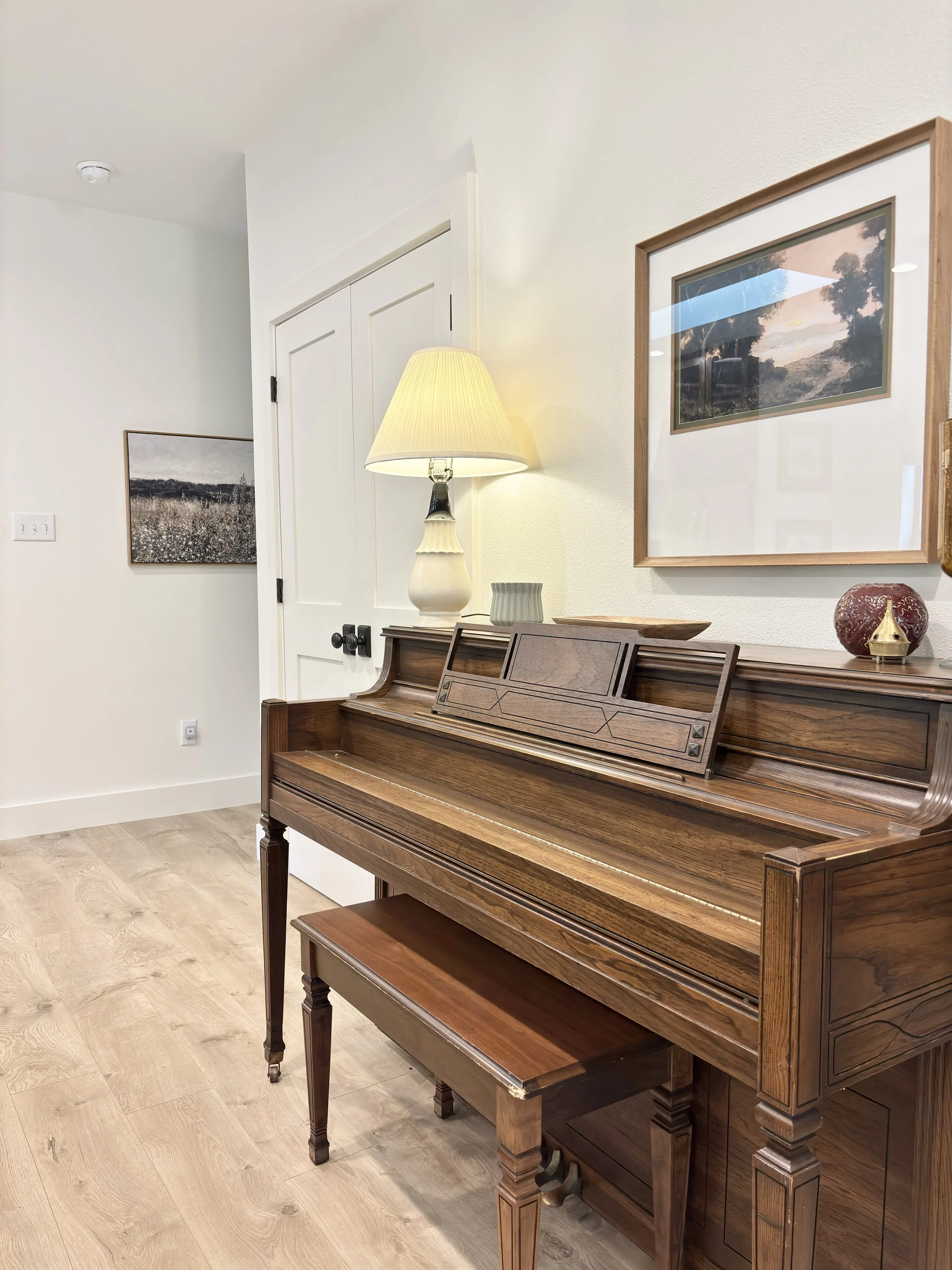 A wooden piano with a matching bench in a room with light-colored walls, framed art, a lamp, and decorative items.