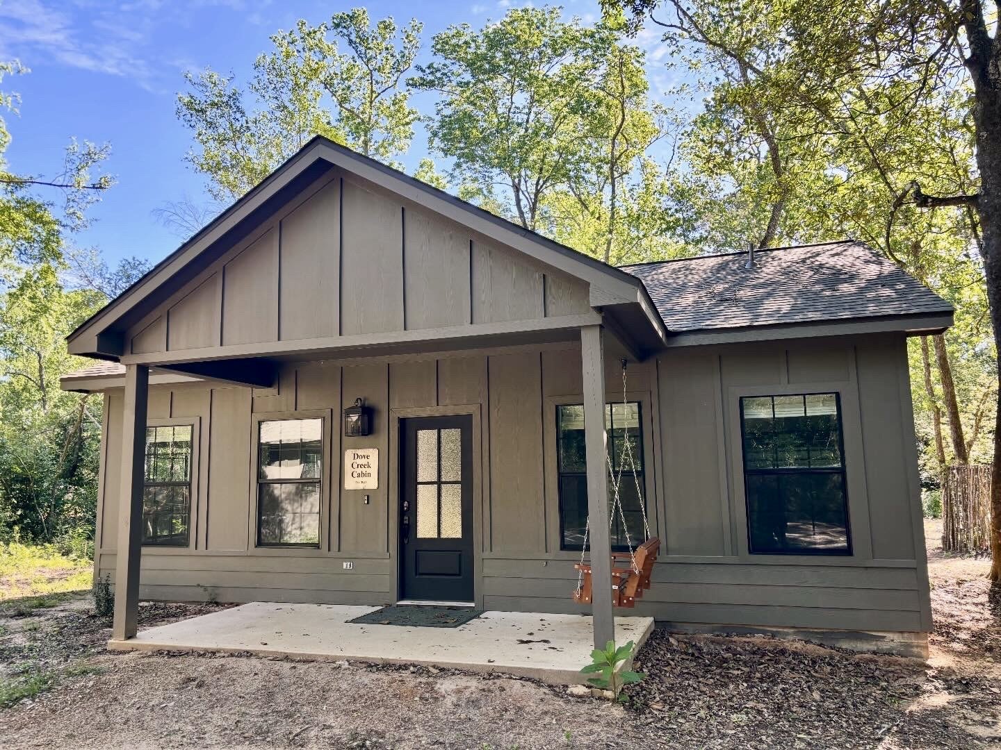 A small gray cabin with a front porch, black door, and four windows, surrounded by trees on a sunny day.