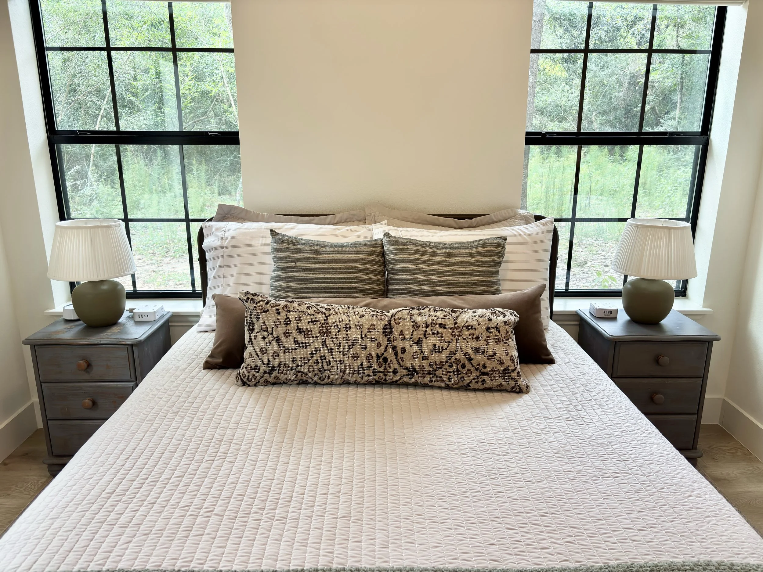 A bedroom with a bed, nightstands on both sides, and large windows showing a view of trees outside.