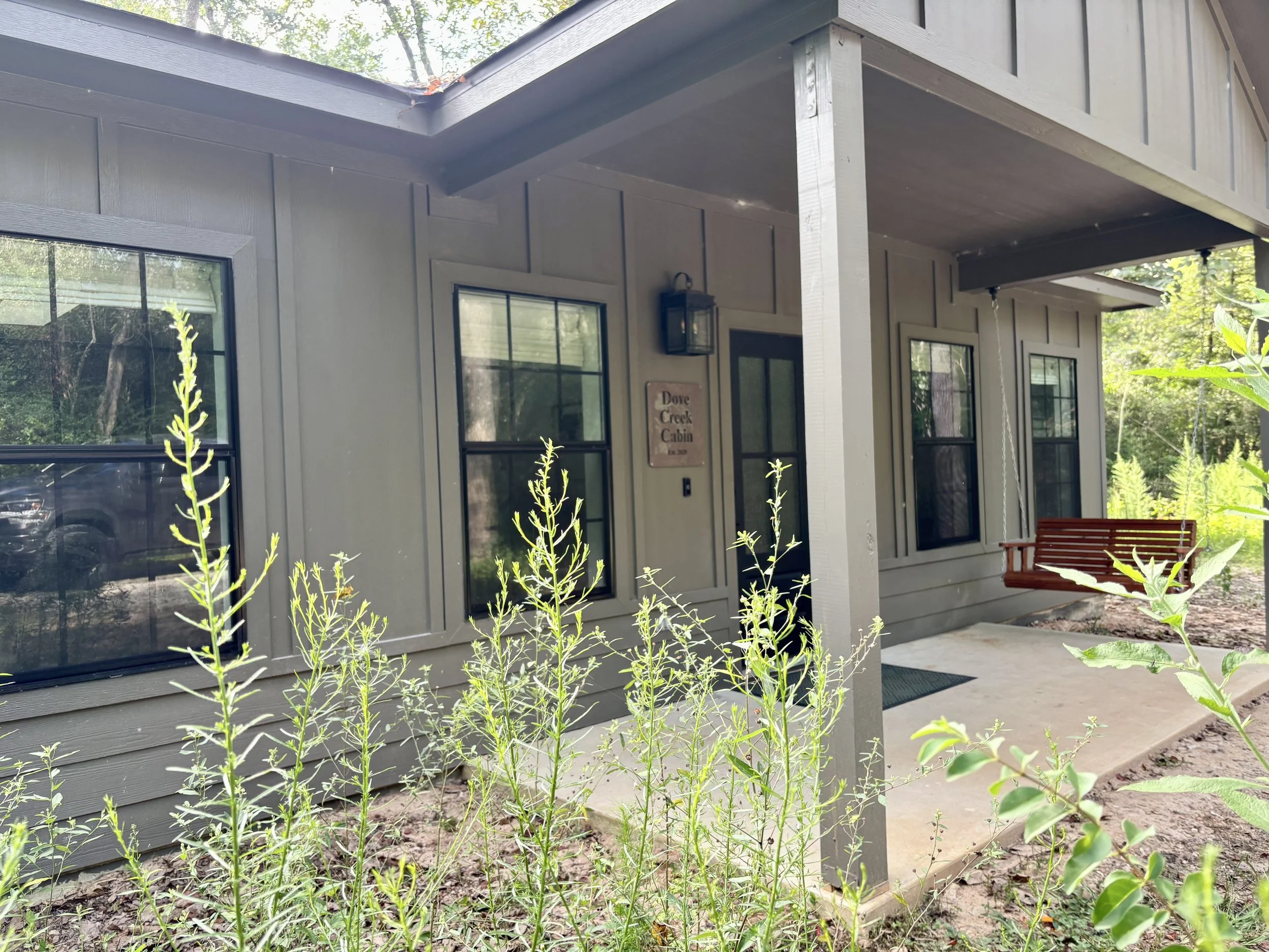 Close-up of the exterior of a modern house with dark gray siding, large black-framed windows, a porch with a swinging bench, and lush greenery around.