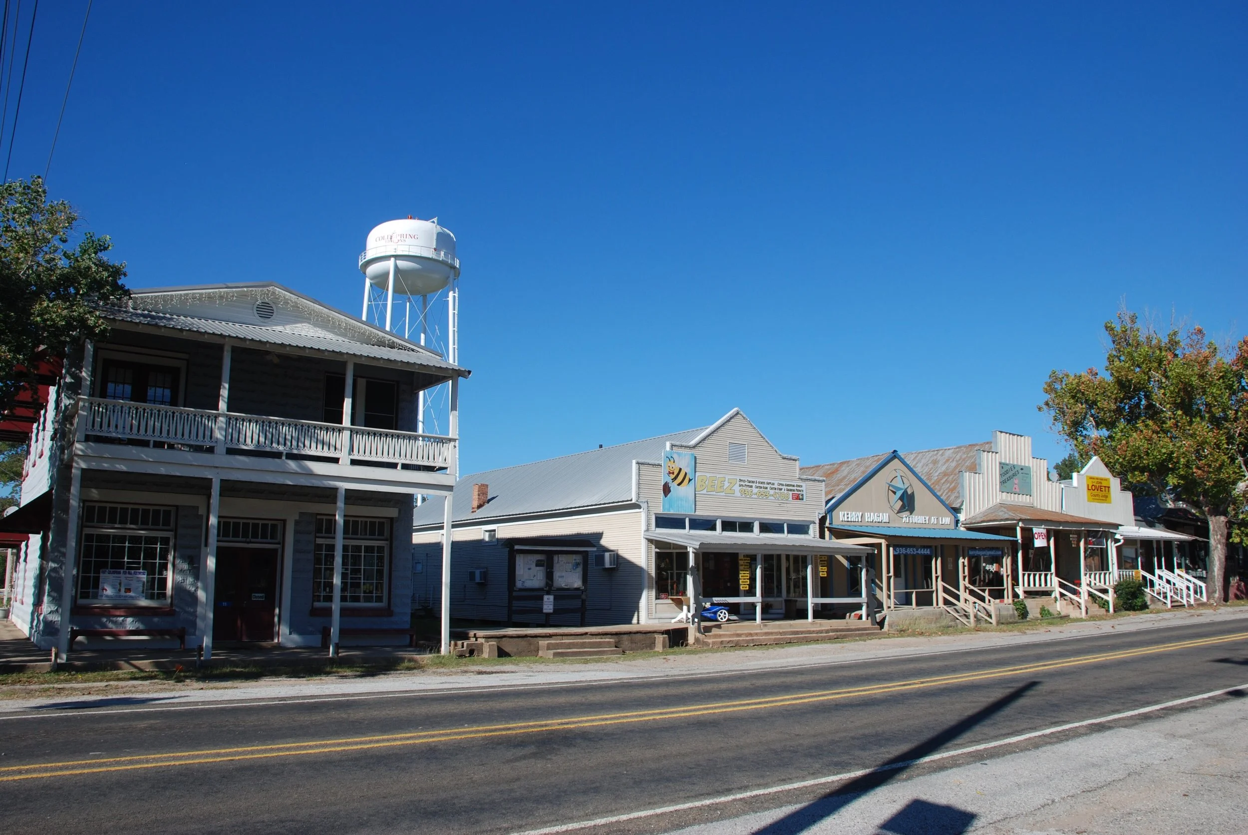 A street view with small shops and businesses including BeeZ, an attorney's office, and other storefronts, some with visible signs. There are trees with autumn foliage on the right and a clear blue sky above. A water tower with the text 'COLLING' is visible in the background.
