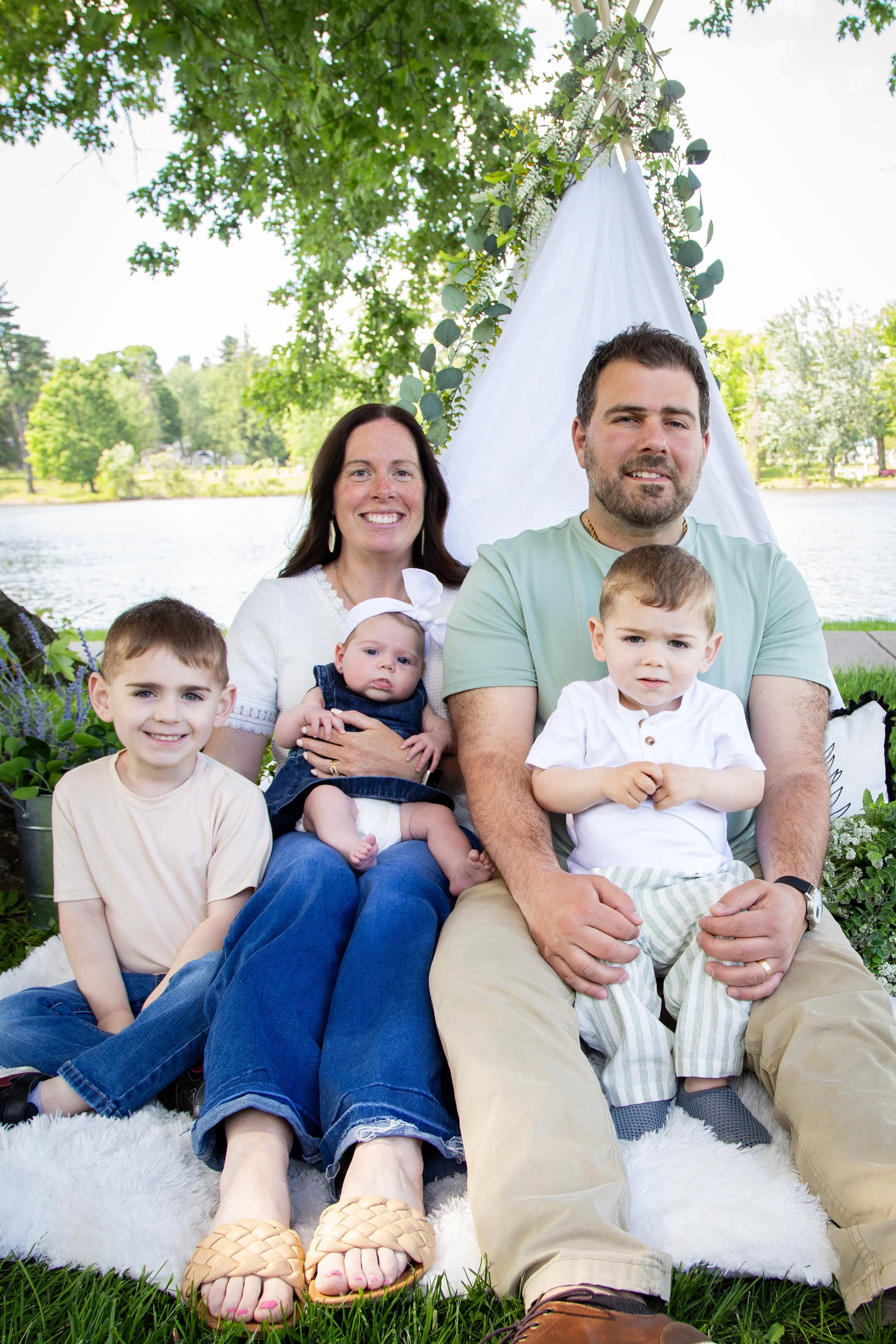 A smiling family of six sitting outdoors by a lake with trees in the background. They are sitting on a white furry rug, with a white fabric canopy decorated with greenery behind them. The family includes two children, a woman, a man, and two babies.