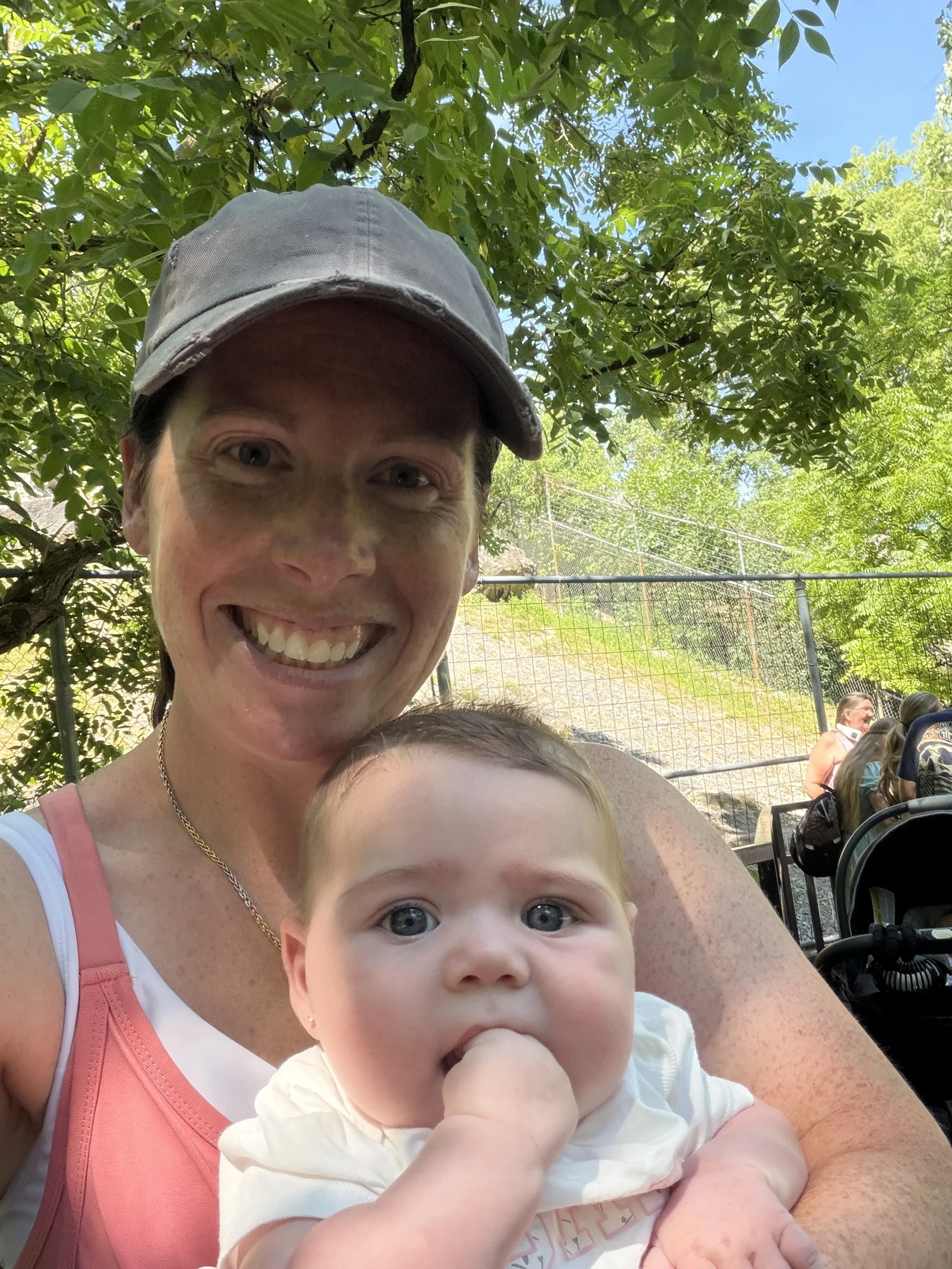 A woman with a gray cap smiling at a camera with a baby in her arms, sitting outdoors among trees. In the background, other people are seated near a fence.